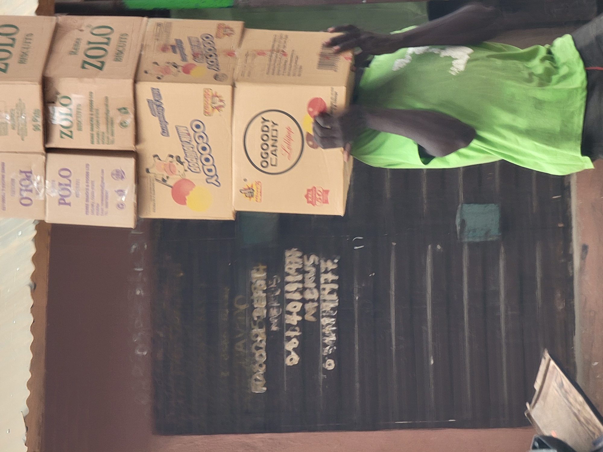 A person wearing a green shirt is holding a stack of boxes, including brands such as "ZOLO Biscuits," "POLO," and "OGOODY Candy Lollipop." The scene likely occurs in a business environment, indicated by the products and the location, which is suggested by the phone number and text partially visible on a wall stating "LAVO Maiburiji." The backdrop is a corrugated metal wall.