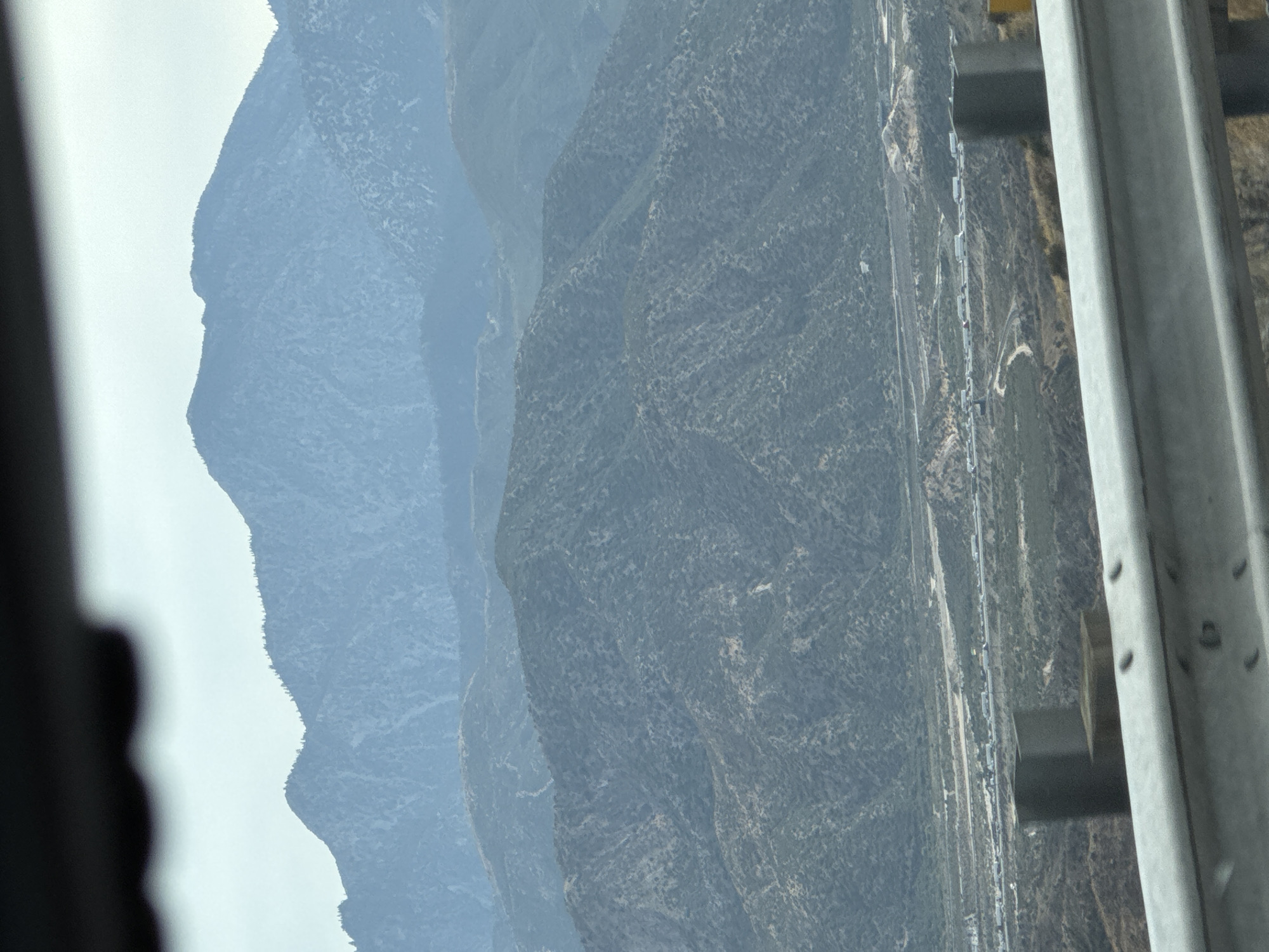 The image displays a wide, mountainous landscape viewed from an elevated perspective, possibly from a vehicle. In the foreground, a metal guardrail runs horizontally across the bottom of the frame, obscuring the immediate view. Beyond the guardrail, the terrain slopes downwards, revealing hills covered in scrubby vegetation. Further in the distance, a highway is visible, with a long line of trucks and other vehicles appearing to be stationary or moving slowly, suggesting traffic congestion. The middle ground consists of rolling, green-brown hills dotted with trees. The background is dominated by several layers of progressively more distant mountains, fading into a hazy, muted blue hue under an overcast sky. The overall scene suggests a rural or semi-rural environment, possibly near a major transportation route. The location of Oak Hills, United States, is provided as context, indicating this likely depicts a view within that area.
