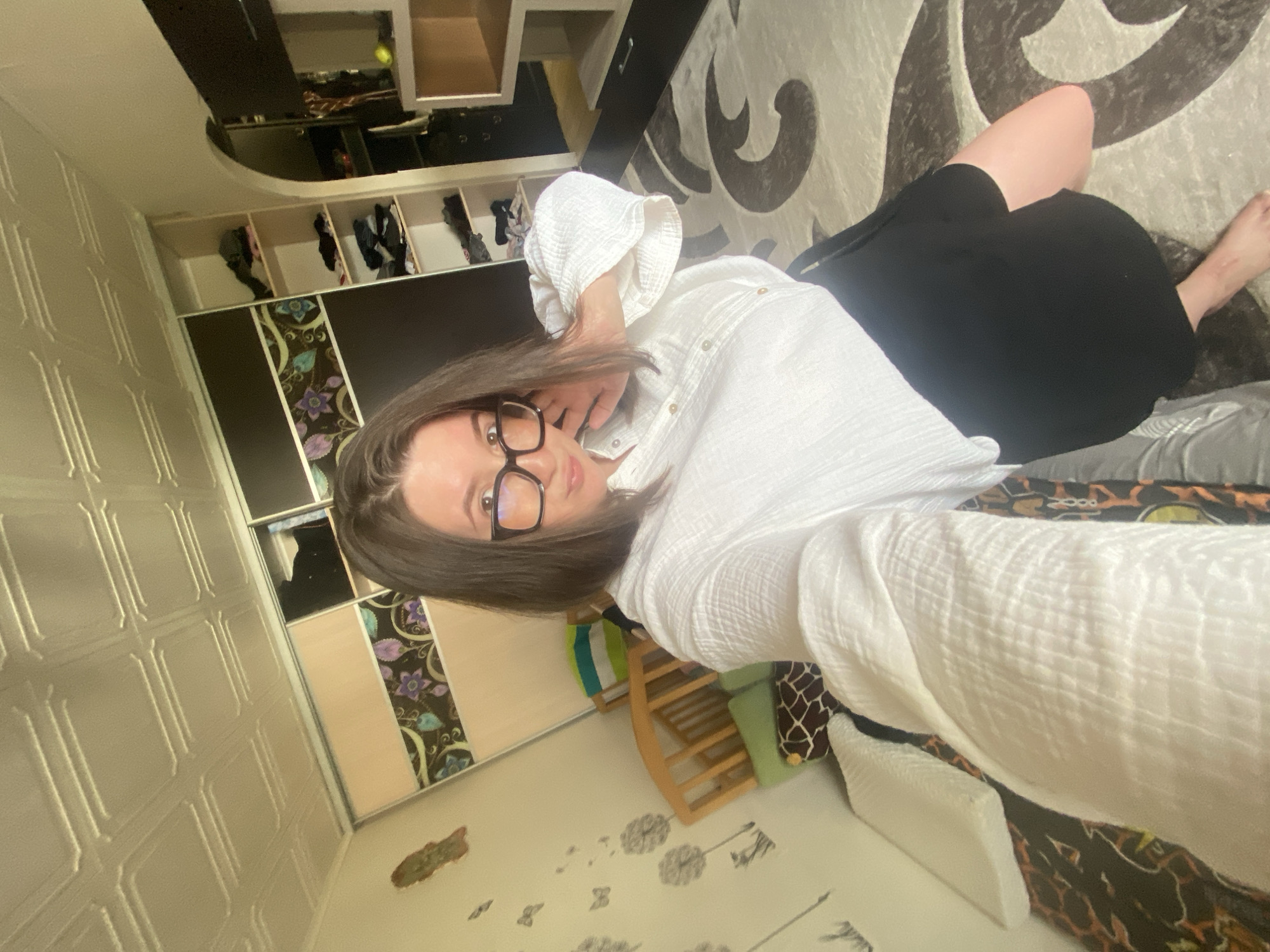A woman with brown hair and glasses is lying on a patterned rug. She is wearing a white button-down shirt and a black skirt. She is indoors in a room with light yellow walls with decorative patterns and a large cabinet with dark wood and floral accents. There is a child's rocking horse and a decorative wall sticker. The location is Chaikovsky, Russia.