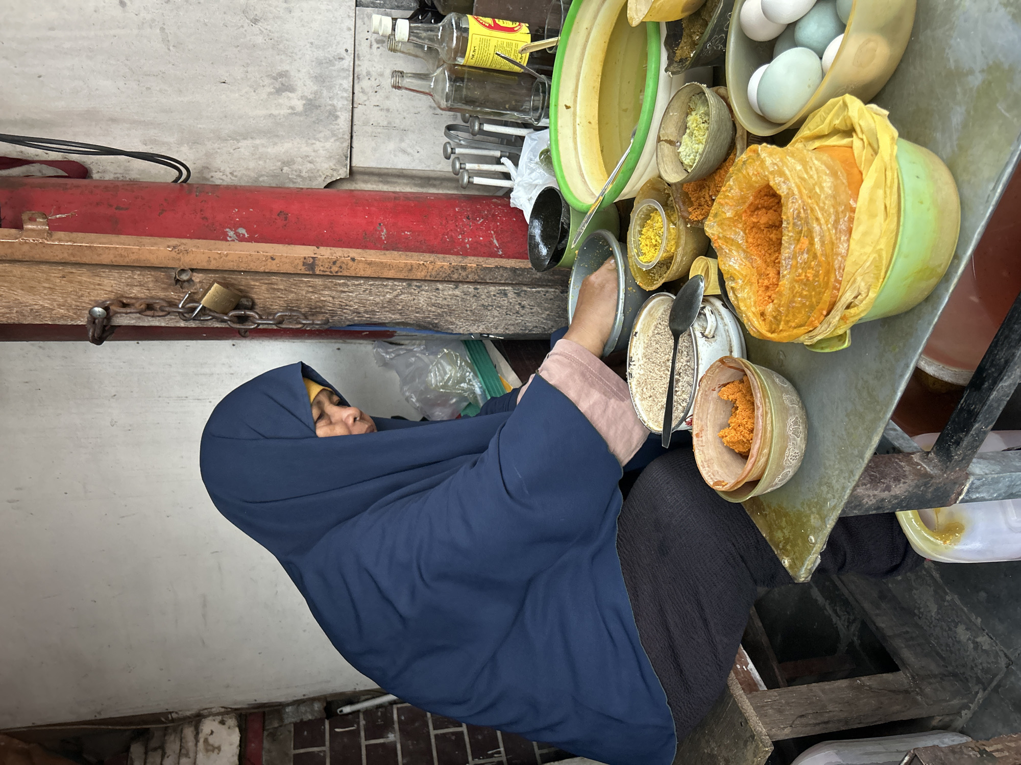 In Yogyakarta, Indonesia, an individual wearing a navy blue hijab and a pink-sleeved garment is seated on a wooden stool, engaged in food preparation. The scene appears to be an outdoor market stall or a small, open-air kitchen. The person's hands are partially obscured by the food items.
On a table in front of the individual are several bowls containing various ingredients. One bowl holds a light brown granular substance, possibly rice or a type of grain, with a spoon resting in it. Another bowl contains a vibrant orange, finely ground spice or powder. There's also a small clear container filled with a bright yellow granular material. A larger green bowl holds a greenish paste or mixture, and next to it, a large pale green bowl is filled with a yellowish liquid. Another bowl, further to the right, contains light green paste. A plastic bag filled with an orange powder is placed on top of a green bowl. In a pale yellow bowl to the far right, several duck eggs are visible, some appearing light blue and others white.
In the background, a red cylindrical object, possibly a propane tank, stands upright, next to wooden planks that are secured with a chain and a padlock. Several glass bottles, including two with yellow labels, are lined up behind the food preparation area, suggesting they contain liquids like cooking oil or sauces. The wall behind the scene is textured and appears weathered. The overall environment suggests a busy, functional workspace where food is being prepared for sale.