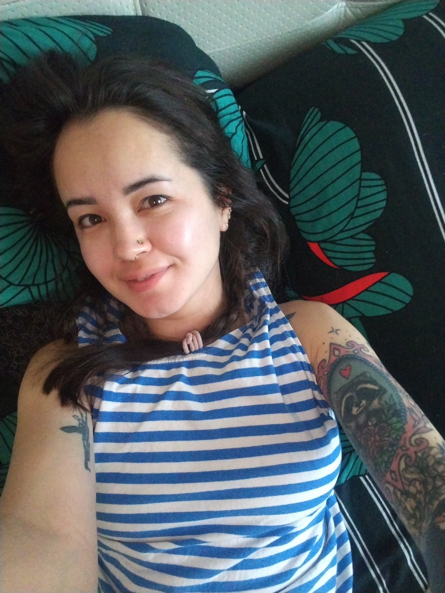 A woman is lying on a bed with a black and green patterned sheet. She has dark hair, a nose ring, and tattoos on her arms. She is wearing a blue and white striped tank top. The location is not discernible as Noviy, Russia.