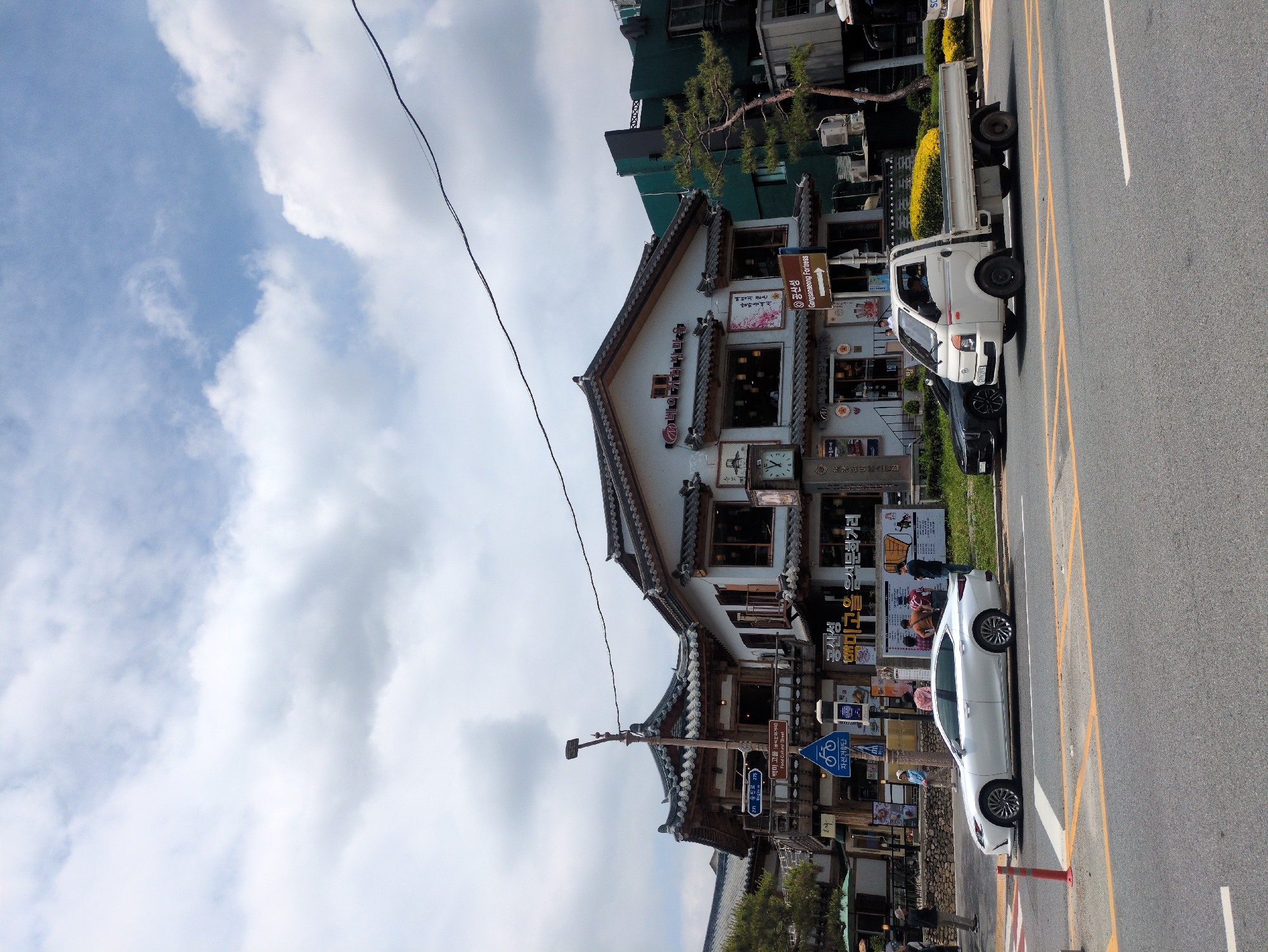The image shows a two-story building in Gongju-si, South Korea, with traditional Korean architectural elements, including a tiled roof. The building faces a street with a white car and a white truck parked on the roadside. Several signs are visible, including directions to Gongsanseong Fortress and information about the Baekmi Goeul Food Cultural Street. Power lines are visible against a blue sky with clouds. People are present near the base of the building.