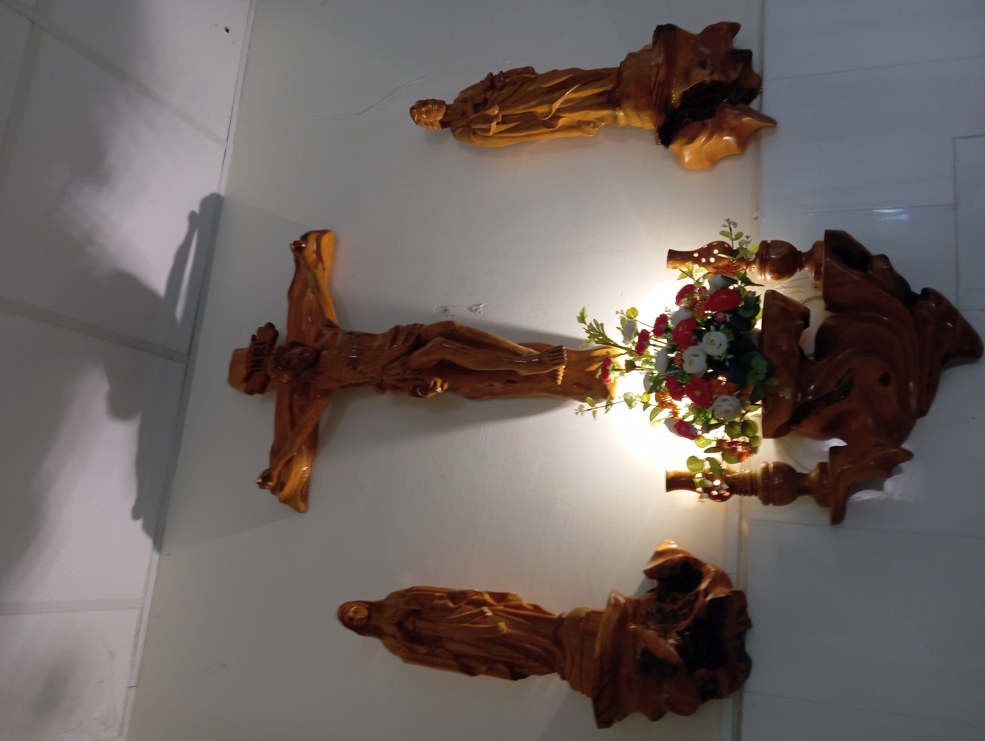 The image depicts a religious altar display, likely within a church or home shrine, in Nha Trang, Vietnam. The central focus is a wooden crucifix depicting Jesus Christ. Flanking the crucifix are two wooden statues, one on the left representing the Virgin Mary and another on the right of an unidentified male saint. Below the crucifix, a decorative wooden shelf holds a vase of artificial flowers and a small wooden lamp. The altar is mounted on a white wall, with shadows cast from a light source above, possibly a ceiling light or a window. The overall scene is one of reverence and devotion.