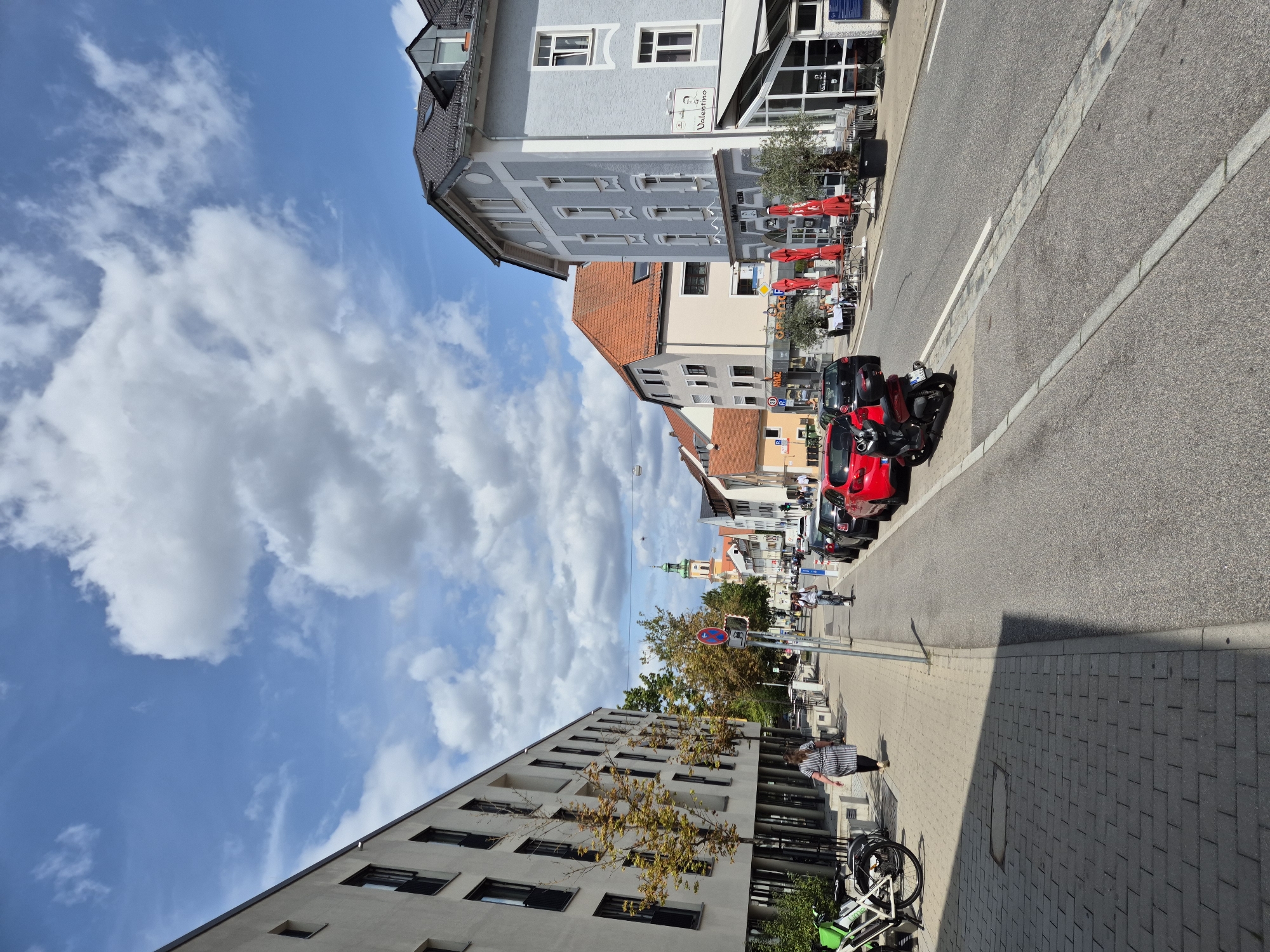 This is a street view of the city of Regensburg, Germany, on a sunny day with scattered clouds. The image captures a street lined with buildings on both sides, a sidewalk, a road with parked cars and a scooter, and a church tower in the distance.
On the left side of the street, a modern, multi-story building with many windows and balconies is prominent. In front of this building, there is a sidewalk with trees and benches. A woman in a striped dress is walking away from the camera on the sidewalk. Several bicycles are parked in a bike rack.
The right side of the street features a row of buildings with different architectural styles and facades. Some of these buildings appear to be commercial establishments, with outdoor seating areas and umbrellas set up in front of them. A light blue building with a gray roof and white trim is particularly noticeable. A sign for "Valentino" is visible on this building, suggesting a restaurant or cafe.
The road itself has two lanes and is lined with parked cars, including a red car and a dark-colored car. A red scooter is parked on the side of the road. Further down the street, a person is riding a bicycle. In the background, the distinct green spire of a church tower rises above the other buildings, a landmark in Regensburg. The sky above is a bright blue with fluffy white clouds.