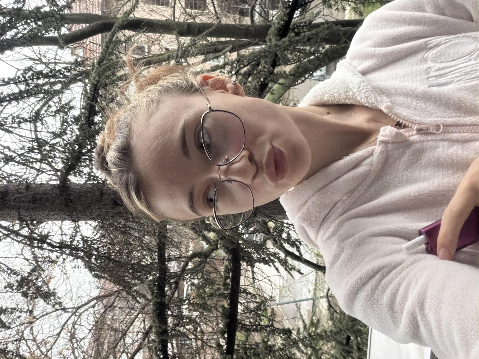 A woman with blond hair styled in a bun, wearing glasses and a pink zip-up hoodie, is holding a pink electronic cigarette device with a white cigarette inserted. She is positioned in front of trees with bare branches. The presence of buildings and trees suggest an urban location, which may be Plovdiv, Bulgaria.