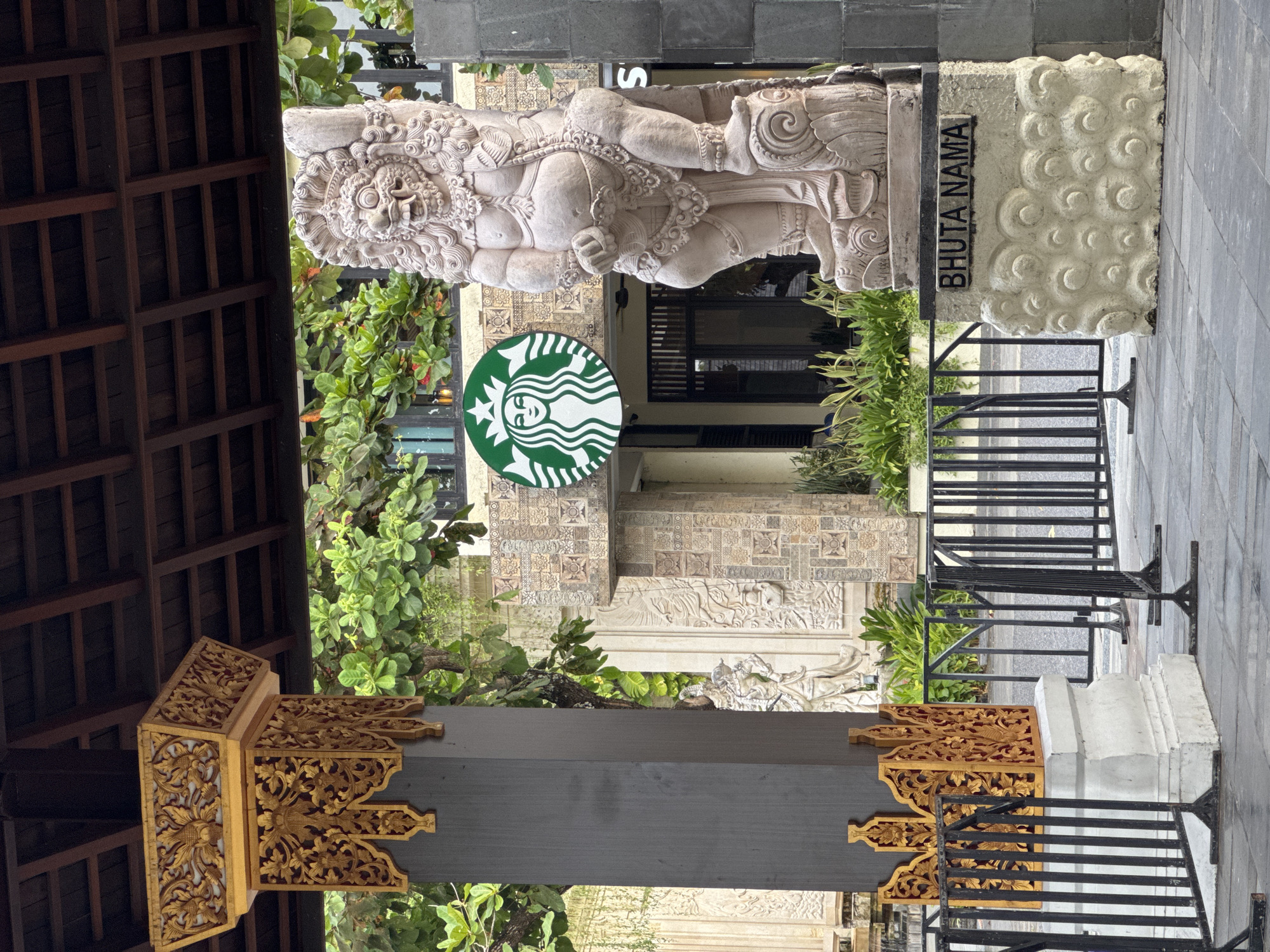 The image shows a Starbucks store in Kuta Selatan, Indonesia. The store entrance is adorned with Balinese statues and carvings. A stone statue of a deity stands above the entrance, and a sign reads "BHUTA NAMA" next to it. The Starbucks logo is prominently displayed on the building facade. The foreground includes decorative wooden carvings with a gold-colored floral pattern. Black metal railings are also visible. Green plants are seen around the building and statues. The ground is paved with gray stone.