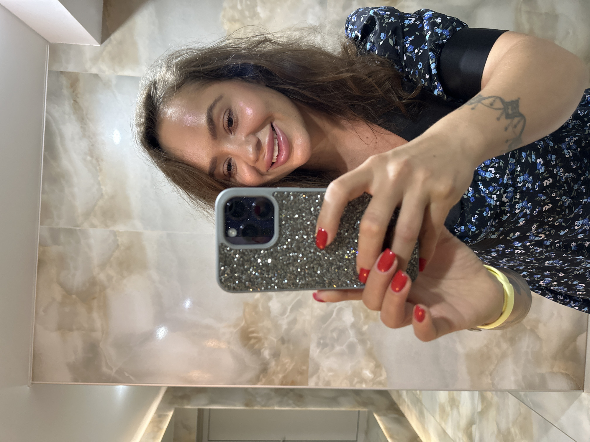 A woman with brown hair and red painted fingernails is taking a selfie in a bathroom. She is smiling and looking at the camera. She is wearing a black dress with blue flowers and has a tattoo on her left arm. The bathroom walls are tiled with a marbled pattern and there is a mirror reflecting more of the room. The image is taken from the chest up.