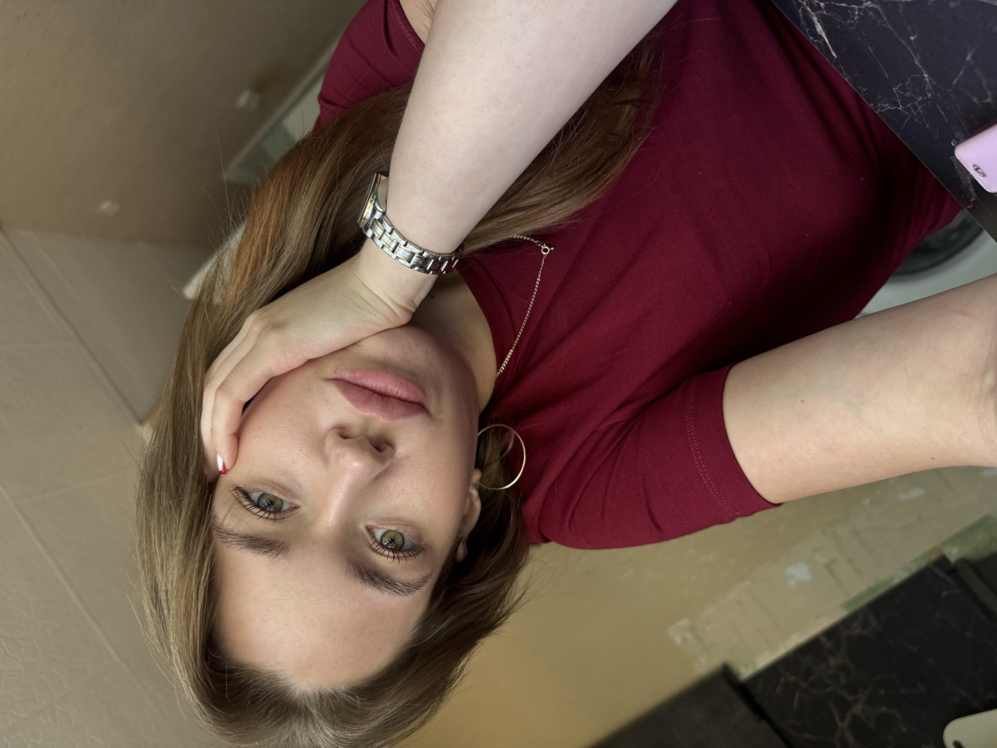 A close-up, rotated portrait shows a young woman with long, light brown hair and green eyes. She is wearing a dark red t-shirt and has a silver watch on her left wrist. She is resting her head on her hand, with her fingers touching her forehead and cheek. A thin gold chain with a circular pendant is visible around her neck, as well as a gold hoop earring in her left ear. Her makeup appears natural, with emphasis on her eyes. The background is out of focus, but suggests an indoor setting with beige walls and a dark countertop. The lighting is soft, indicating it might be daytime, possibly indoors with natural light or soft artificial lighting. There is no visible text in the image.
