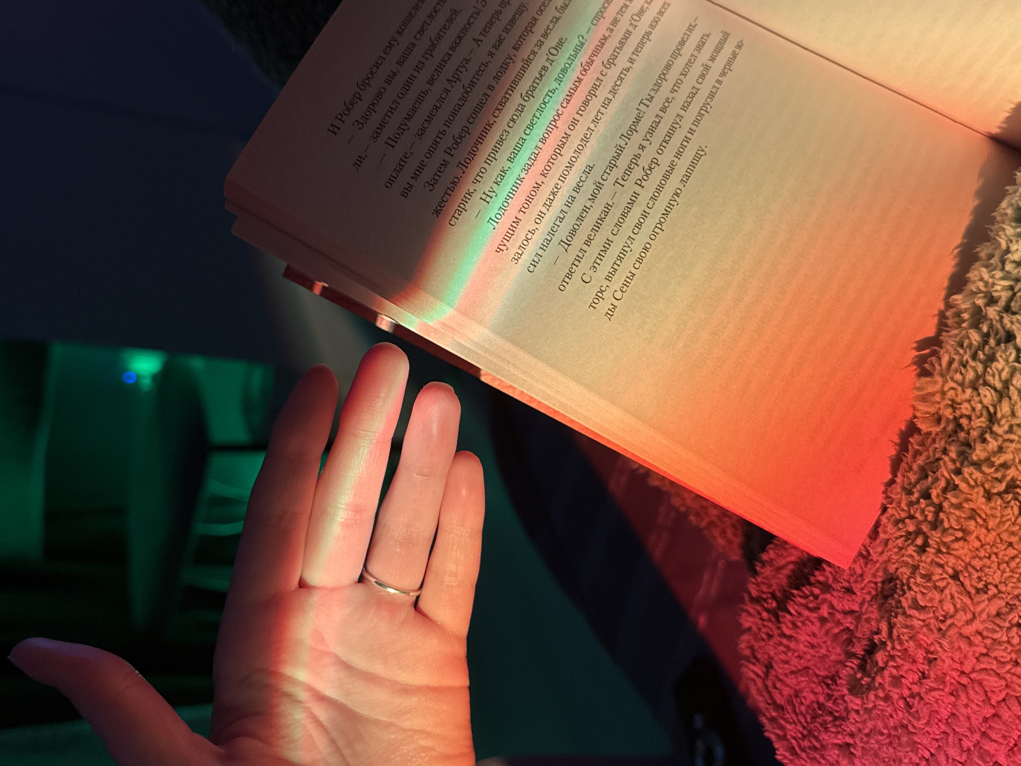 A close-up shot captures a human hand adorned with a simple silver-colored ring on its ring finger, positioned near an open book. The scene is illuminated by vibrant artificial lighting, casting distinct hues across the composition. The left side of the frame, including part of the hand and the background, is bathed in a deep green light, while the right side, encompassing the open book and the remainder of the hand, glows with a warm orange-red. A gradual color transition, resembling a rainbow, is visible where these lights meet on the book's pages and the palm of the hand.
The open book rests on a soft, fuzzy, orange-red textile, likely a blanket, suggesting a cozy, indoor reading environment, possibly in the evening or at night. The book's pages display text in Cyrillic script. The visible passage describes a narrative involving characters named "Робер" (Rober), "Артуа" (Artois), and "Лорме" (Lorme), engaged in dialogue about money and satisfaction, with mentions of a boatman and physical descriptions. Phrases like "И Робер бросил ему кошелев" (And Rober threw him a purse), "Здорово вы, ваша светлост" (You're great, your highness), and "Ну как, ваша светлость, довольны?" (Well, your highness, are you satisfied?) are discernible. The hand's placement suggests the person is actively reading or about to turn a page.