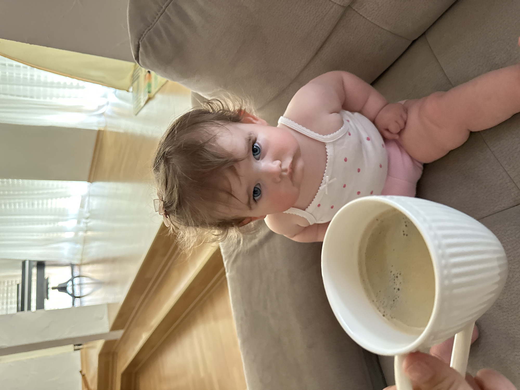 A baby is lying on a tan-colored couch. The baby has fair skin, blue eyes, and light brown hair with a small pink hair clip. The baby is wearing a white top with small pink polka dots and a white bow. The baby is looking directly at the camera. In the foreground is a white ribbed mug filled with coffee held by a hand. The background shows a wooden floor, a window with white blinds, and a partially visible curtain. The location is not provided in the image and cannot be determined.