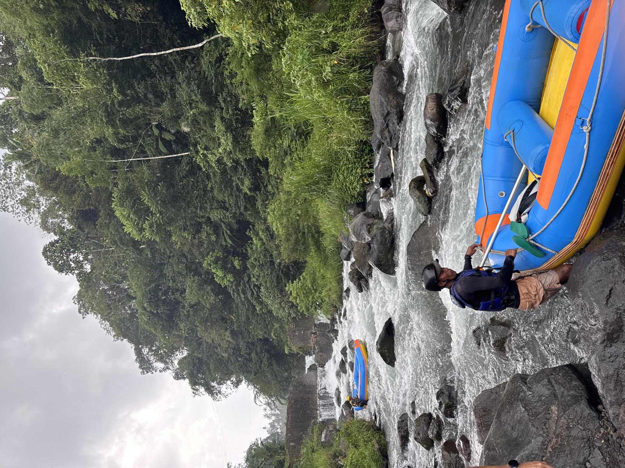 A man in a helmet and life vest stands on rocks in a fast-flowing river, holding onto the side of a blue and yellow inflatable raft. The river is bordered by lush green vegetation and trees. In the background, another raft with people is visible in the river. The sky is overcast. The location is Selat, Indonesia.