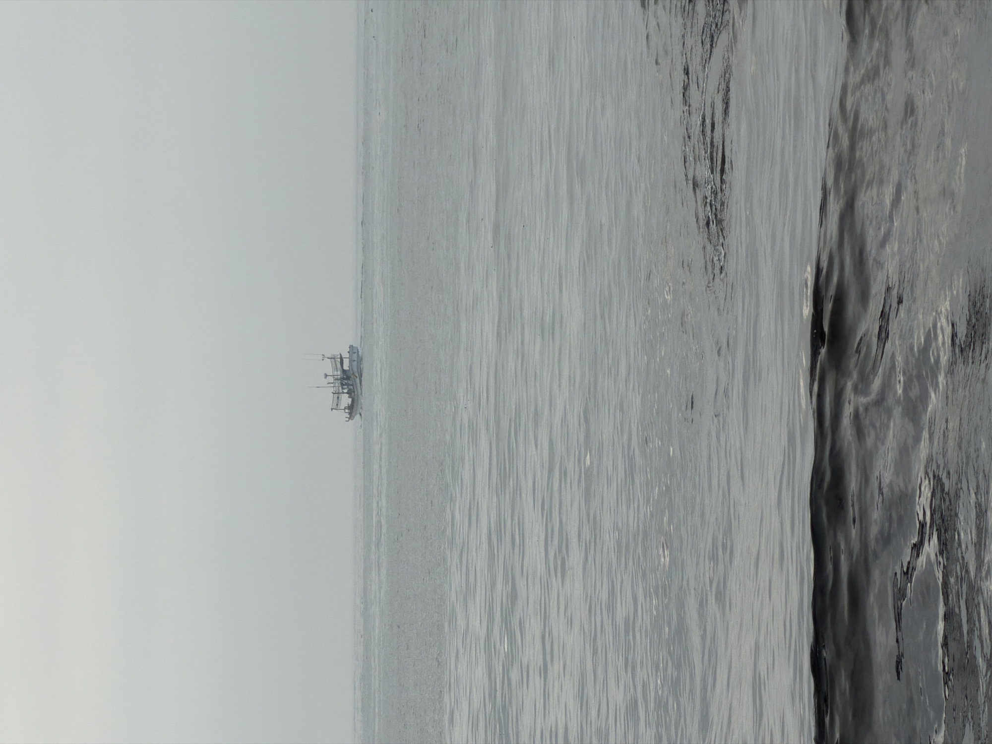 A fishing boat is navigating the calm, gray waters of the sea. The sky is overcast, creating a muted, diffused light. The boat, small in the frame, is positioned towards the right side of the image, appearing as a white silhouette against the hazy horizon. The foreground shows the textured surface of the water with gentle ripples. The scene evokes a sense of quiet solitude and the vastness of the ocean. This location is not specific to Jeju city.