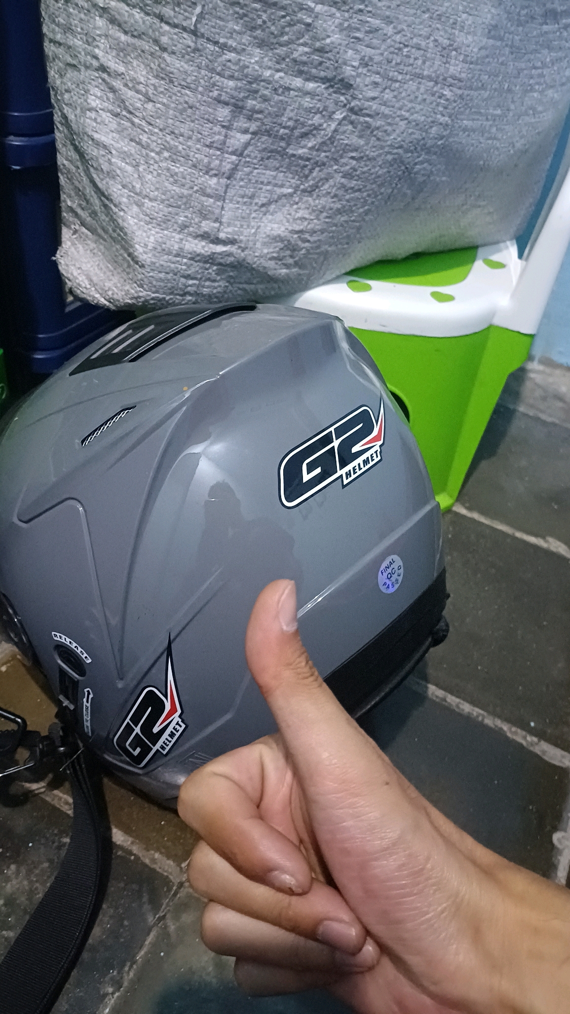 A first-person perspective shot shows a person's right hand giving a thumbs-up gesture, with the thumb and part of the index finger in focus, in front of a gray motorcycle helmet. The helmet has a sticker with the text "G2 HELMET" and another small circular sticker with "FINAL QC" and some numbers. The helmet rests on a tiled floor, next to a green and white plastic container and a large, textured gray bag. The environment suggests an indoor or sheltered outdoor space, possibly a storage area or garage. There is no indication of the city of Kowangan, Indonesia, in the image.