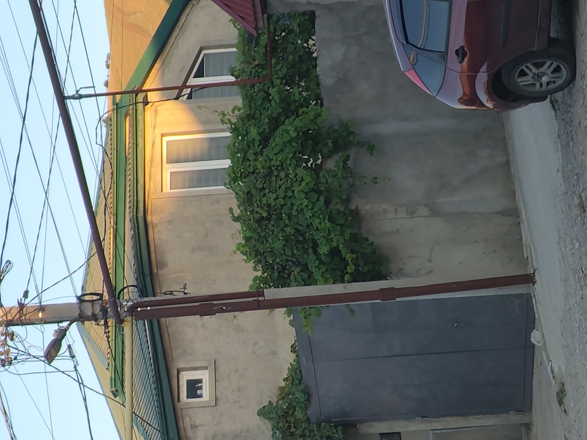 The image depicts a residential property in Leninkent, Russia, characterized by a two-story house with a green corrugated roof and light-colored walls. A large, dark gray metal gate is visible on the left side of the property, partially obscured by lush green foliage. The house features multiple windows, including a small square window on the upper floor and larger rectangular windows with white frames on the second level. A street lamp is mounted on a utility pole positioned in front of the house, surrounded by a complex network of overhead wires. A portion of a dark red car is visible in the lower right corner of the frame, parked on the street adjacent to a concrete wall. The surrounding environment appears to be a moderately developed urban or semi-urban area.