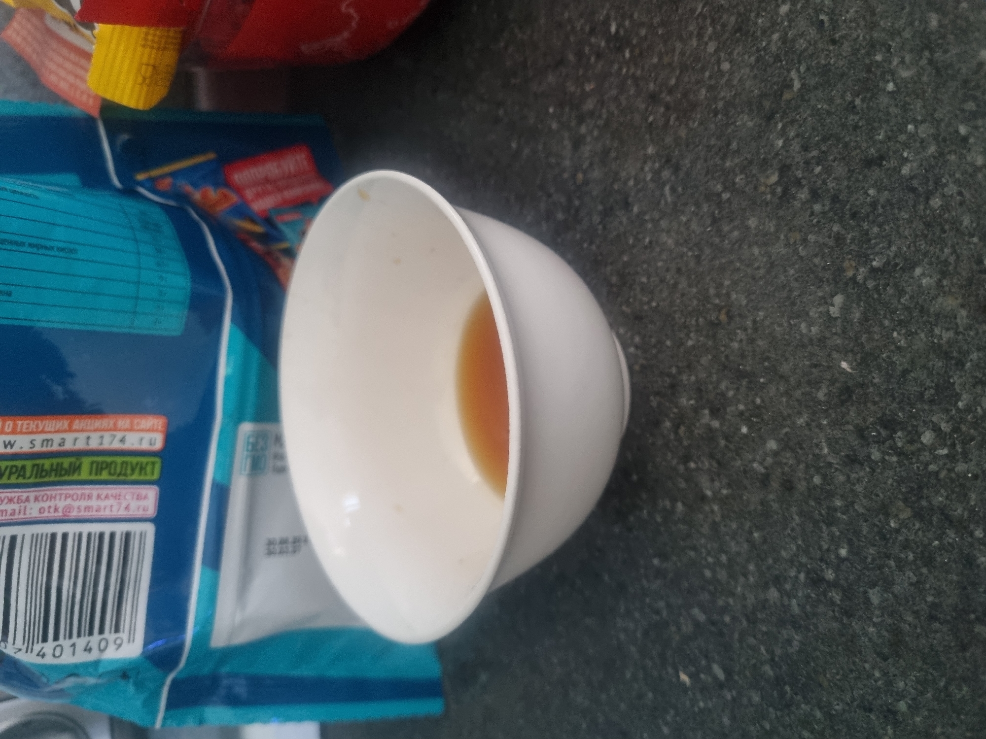 The image shows a white ceramic bowl containing a small amount of amber-colored liquid, likely tea or coffee, placed on a speckled dark gray countertop. In the background, a blue and white packaging with Cyrillic text is visible. The packaging has a barcode and nutritional information printed on it. The scene appears to be indoors, possibly in a kitchen or dining area. There is no discernible location in the city of Samara, Russia, within the image itself. No people are present in the frame.
