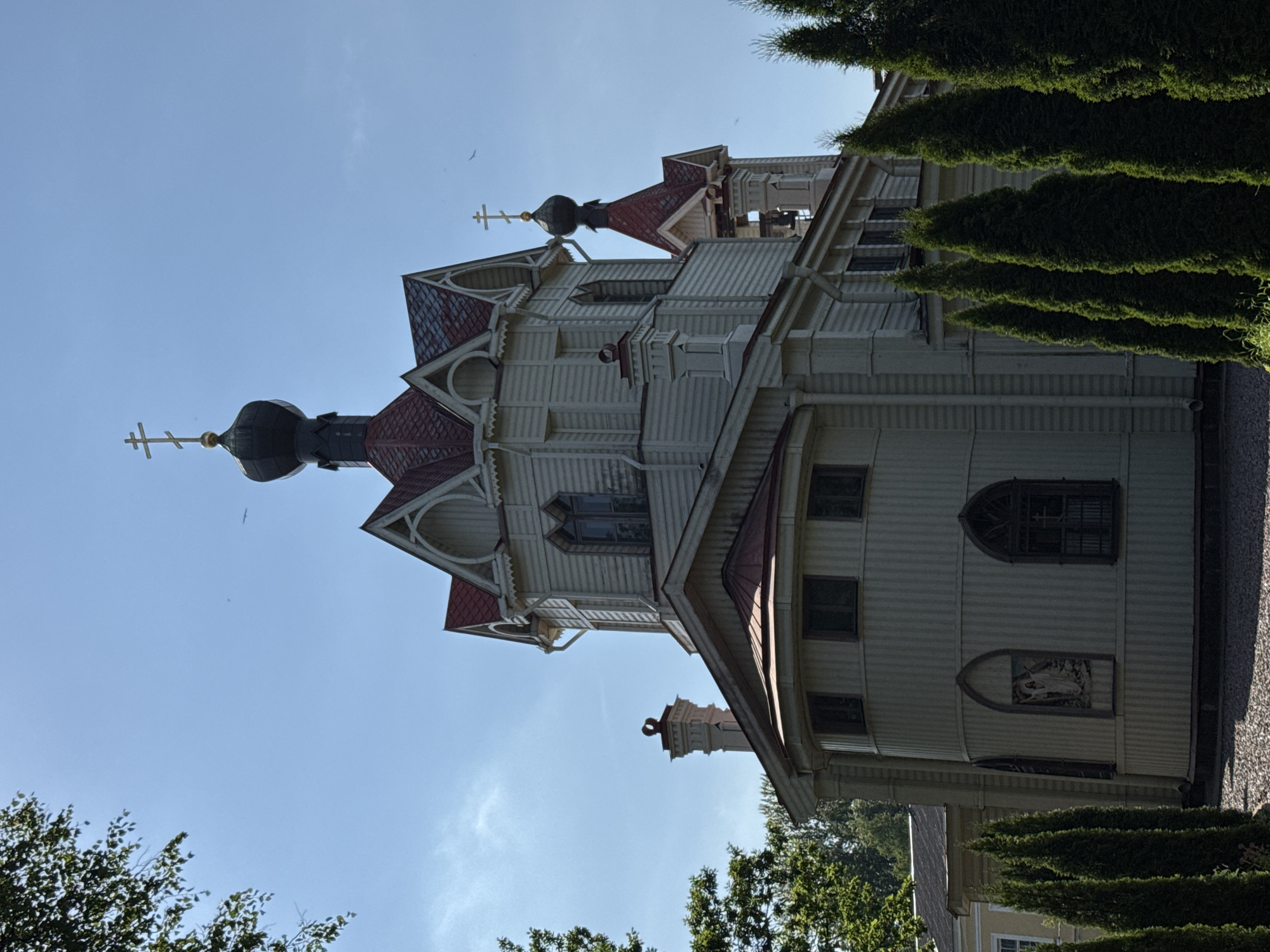 This is a media file of an outdoor scene captured in Lomonosov, Russia, with the camera oriented vertically. The primary subject is a beautiful, white wooden church with a red roof, featuring intricate architectural details such as decorative gables, pointed spires topped with crosses, and arched windows. A dark, bulbous dome crowns one of the spires, also adorned with a cross. The church is partially obscured by lush green coniferous trees on the right and some deciduous trees and branches in the lower left and bottom of the frame. The sky is clear blue with a few wispy clouds, indicating a bright, sunny day, likely in the late afternoon or early evening given the soft lighting. There is no visible text in the image. The scene evokes a sense of tranquility and historical charm.