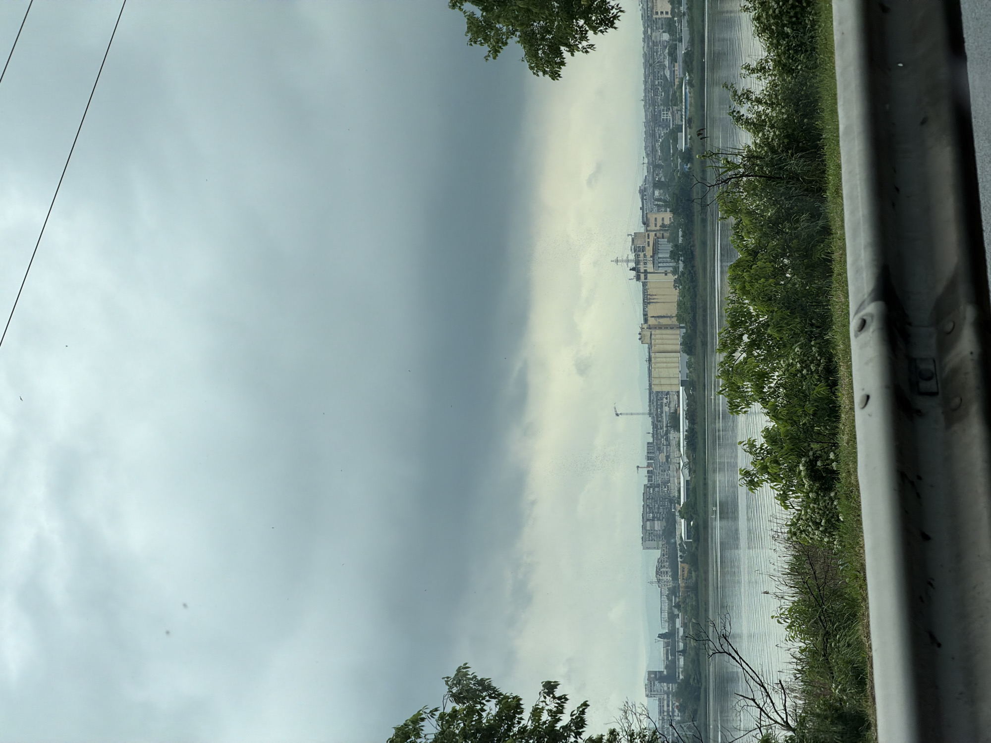 The image shows a cityscape likely in Burgas, Bulgaria, viewed from an elevated position, possibly a bridge or high point along the coast. The sky is overcast with dark, stormy clouds on the left side transitioning to lighter, grey clouds towards the right. The cityscape includes industrial structures and residential buildings along the water. Tall industrial silos are prominent. Water separates the viewpoint from the cityscape. Vegetation is visible in the foreground. A metal guardrail is present.