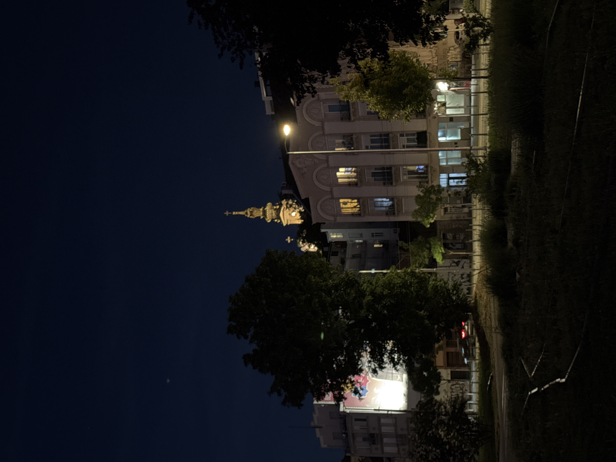 At night, the image shows a cityscape with a street and buildings. A prominent church steeple is visible. The location appears to be Belgrade, Serbia, given the architectural style. A streetlamp illuminates a building. There's vegetation to the right. A dark sky dominates the left portion.