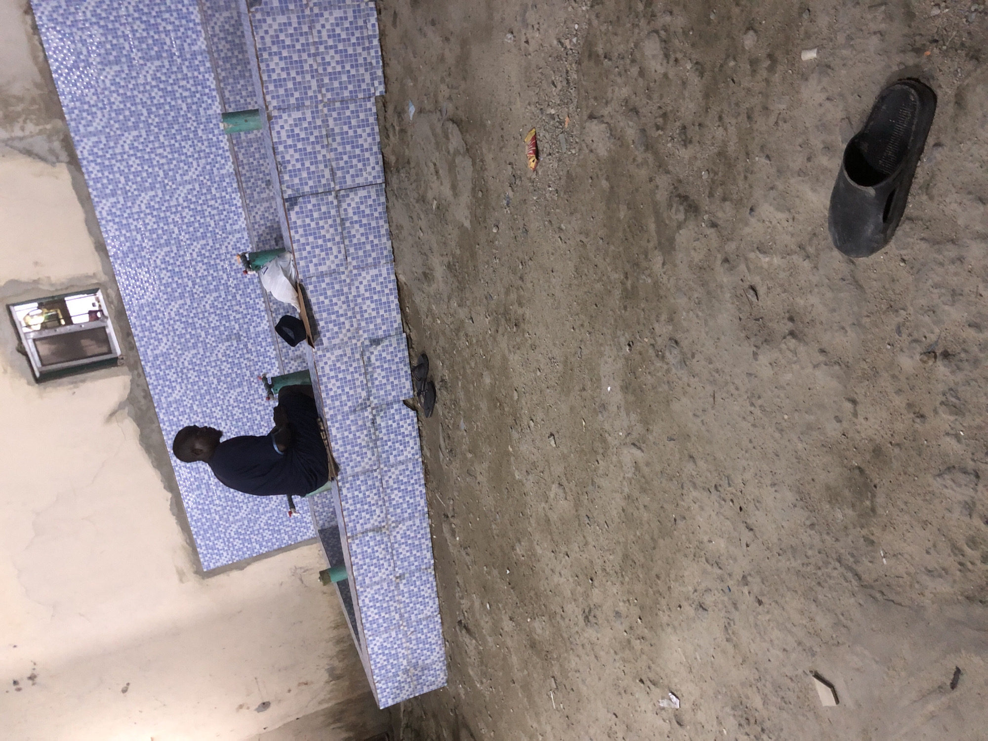 In Ado, Nigeria, a man sits on the edge of a tiled basin, which appears to be part of a public ablution facility. He is wearing a dark blue traditional garment and is facing away from the camera, looking towards a row of green plastic taps. In front of the taps, there are several items on the basin's edge, including a black cap and a white plastic bag. The facility is constructed with blue and white square tiles, and the surrounding area is composed of dry, dusty earth. A small, dirty window is visible on the upper wall. Further from the basin, on the ground, a black slipper lies discarded. The scene suggests a functional, albeit somewhat rough, urban environment.