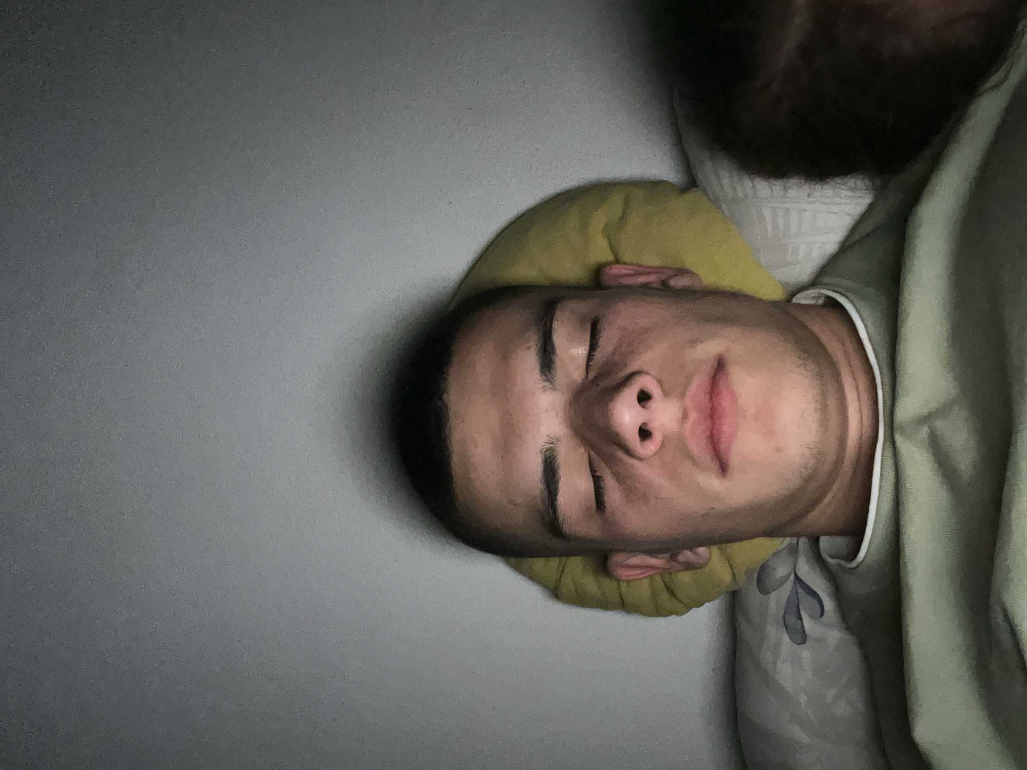A medium shot shows a man sleeping in a bed. He is lying on his back with his eyes closed. He is fair-skinned with dark hair and eyebrows. He is resting his head on a yellow pillow. Part of another person's head with dark brown hair and a beard is visible in the upper right corner of the image. The man is covered with a light green blanket over patterned sheets. The background is a light gray wall. The location and time of day are unknown, but the lighting suggests it is indoors and possibly at night. There is no evidence to indicate the location is Karavelovo, Bulgaria.