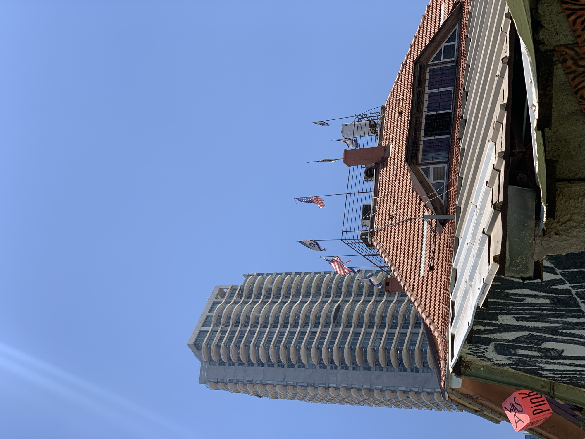 This is an outdoor, eye-level, medium shot captured on a bright, clear day with a vibrant blue sky. The image features a tall, modern apartment building with numerous balconies, situated behind a lower, older building with a red-tiled roof. The older building has a slanted metal roof in the foreground, and graffiti is visible on its side. Several flags are displayed on the rooftop of the older building, including the American flag and an Israeli flag, along with other flags that are difficult to identify. The taller building appears to be a residential high-rise with a contemporary architectural design, characterized by its curved balconies and repetitive facade. The scene suggests an urban environment, likely in Tel-Aviv, Israel, as indicated by the presence of the Israeli flag and the architectural style.