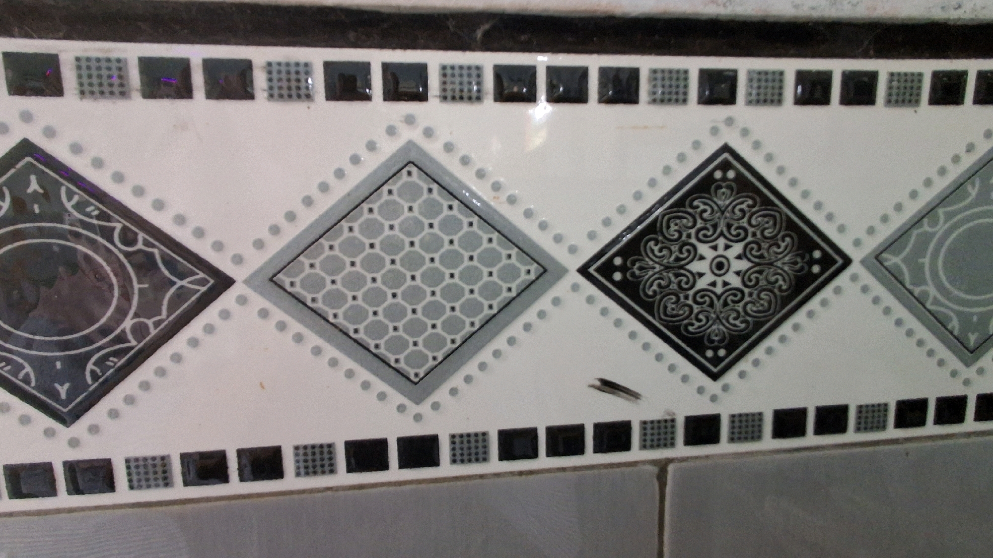 The image shows a decorative tile border, likely in a bathroom or kitchen, in Giriwarno, Indonesia. The border is composed of a series of square tiles with a repeating pattern of dark squares and textured squares. Above this border, there is a row of diamond-shaped tiles, each with a unique black and white or gray and white geometric design. One diamond tile features a complex swirling pattern, while another has a honeycomb-like design. A small insect is visible on the white tile between two diamond patterns. The tiles are set against a white background. The overall impression is of a tiled wall with a decorative accent.