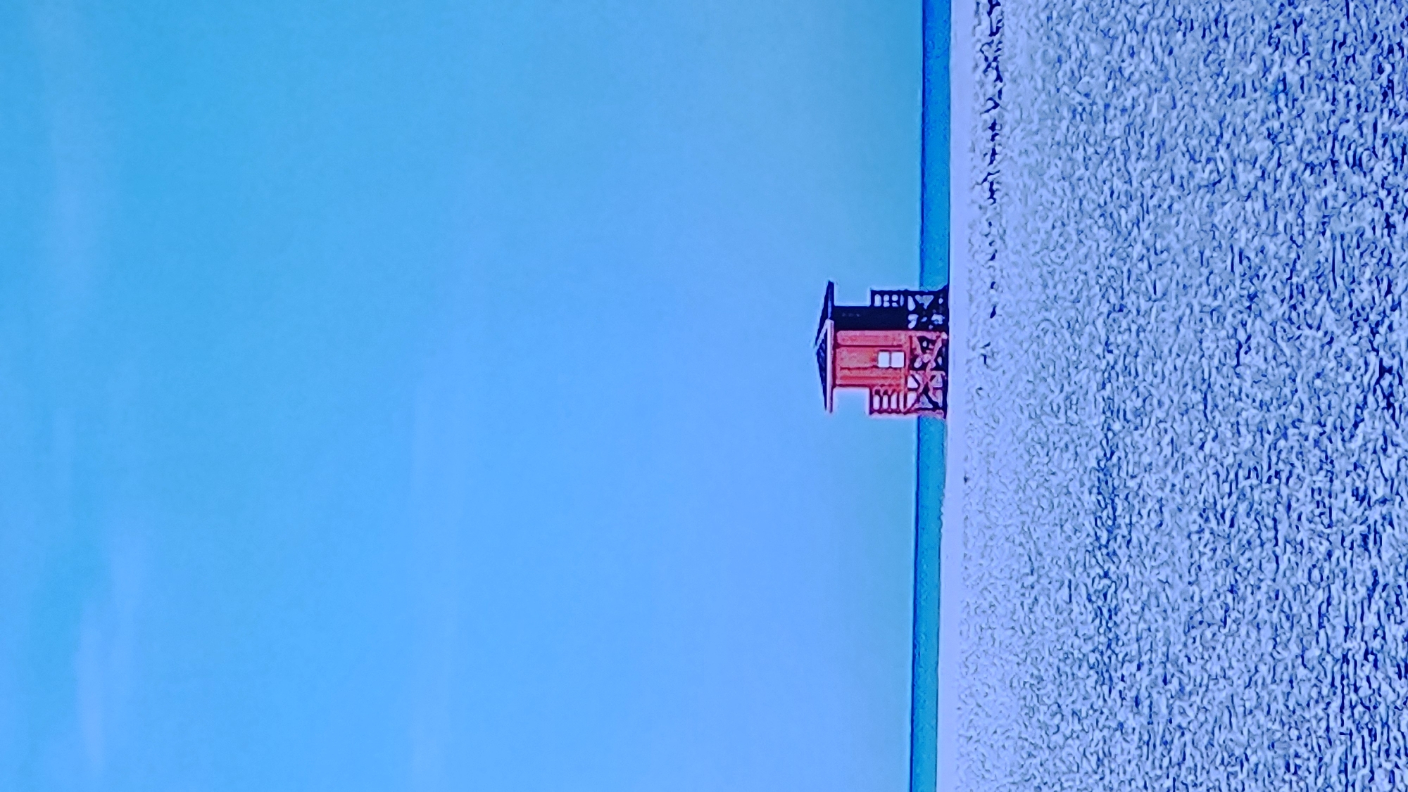 The image depicts a solitary red lifeguard tower on a sandy beach under a clear blue sky. The tower is small, elevated on stilts, and has a peaked roof. It is positioned in the center of the frame, with the vast expanse of sand in the foreground and the calm blue ocean meeting the horizon behind it. The lighting suggests a bright, sunny day. There are no people visible in the scene. The environment is serene and minimalist, emphasizing the isolation of the tower against the natural backdrop.