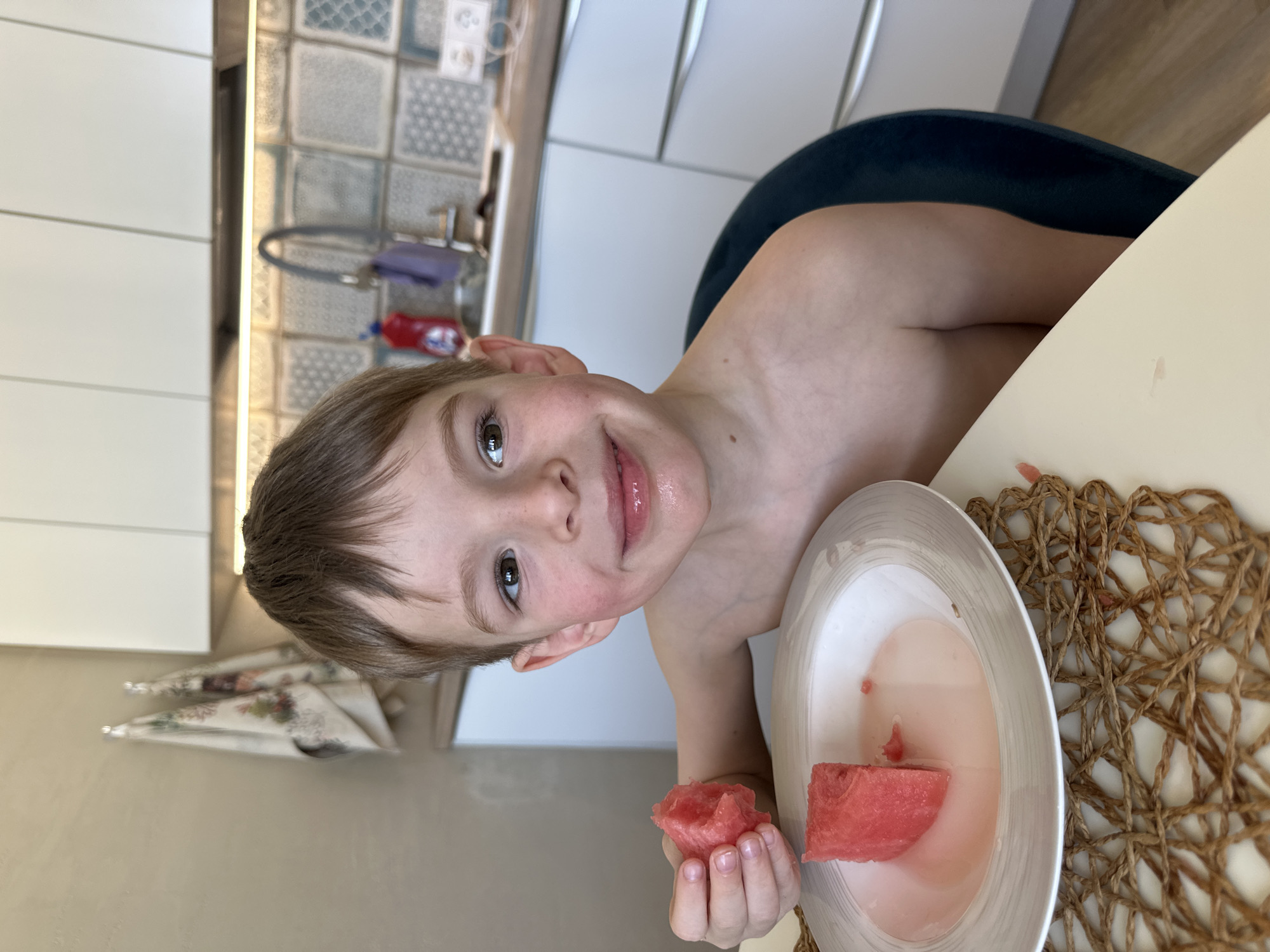 A young, shirtless boy is sitting at a table in what appears to be a kitchen in Chaikovsky, Russia. He is holding a piece of watermelon in his left hand. There's another piece of watermelon in a white plate on the table. The table has a woven placemat. The background reveals a tiled wall and white kitchen cabinets.
