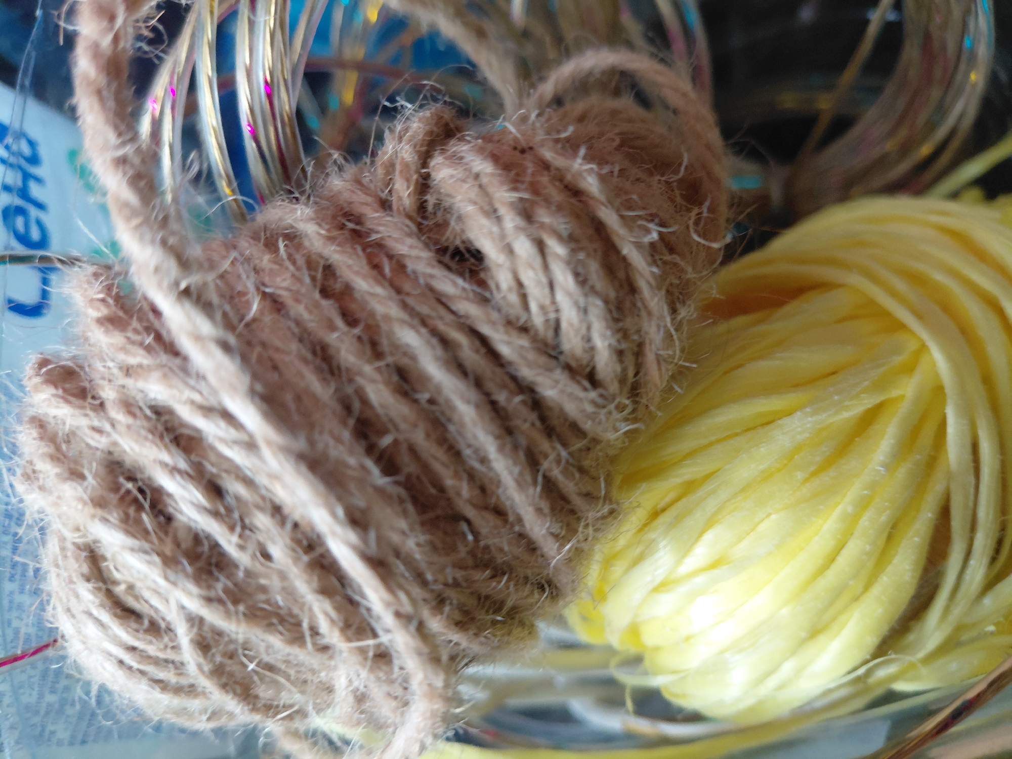 This close-up shot features two bundles of string or yarn, likely craft materials, set against a blurred background. On the left, a rustic-looking bundle of brown, natural-fiber twine, possibly jute, is prominent. Its texture is rough and slightly fuzzy, with individual strands visible. To its right, a vibrant yellow bundle of smooth, somewhat shiny material, possibly raffia or plastic ribbon, is tightly wound, creating distinct, parallel lines.
In the background, translucent tubes or strands with colorful glitter (pink, blue, yellow) are visible, suggesting decorative elements. A blue and white object, possibly a container or package, is partially seen on the left side of the background. On this object, blurred Cyrillic text is discernable, with letters that appear to spell "Лена" (Lena), a common name or part of a brand name.
The image is a still life, focusing on the textures and colors of the different materials. No specific activity is occurring, and no emotions are conveyed. The lighting appears bright, highlighting the distinct qualities of the threads. The location is contextualized as Nadym, Russia, although no specific visual cues within the image confirm this.