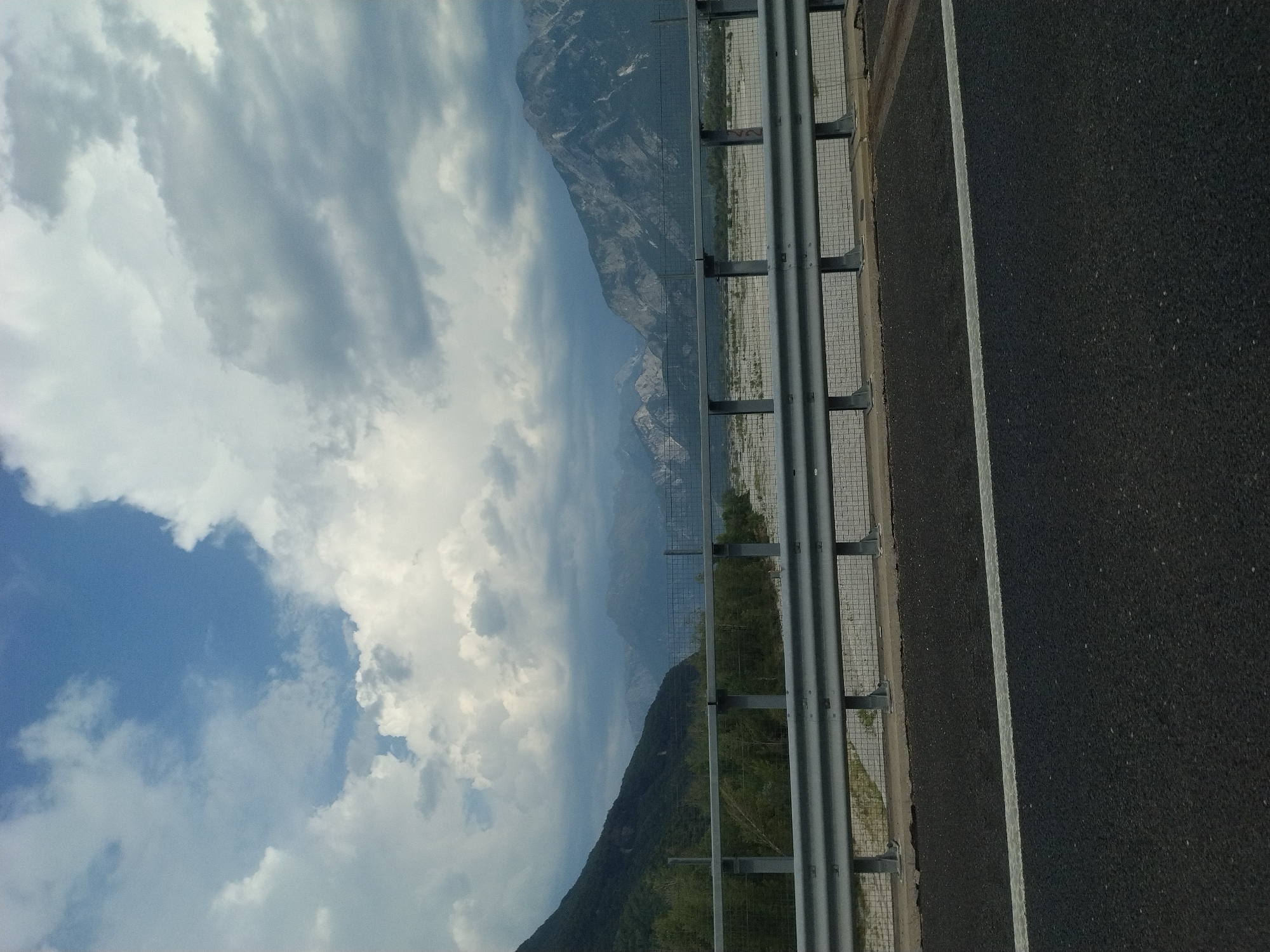 A wide shot captures a scenic mountain vista from a highway, with a guardrail prominently in the foreground. The sky is a dramatic mix of blue and heavy white and grey clouds, suggesting dynamic weather. Below the guardrail, a wide, gravelly riverbed stretches out, bordered by green vegetation and then the imposing, forested slopes of a mountain on the left. In the distance, the rugged, rocky peaks of more mountains are visible, shrouded in atmospheric haze. The highway itself, paved with dark asphalt and marked with a white line, occupies the bottom portion of the frame, indicating the viewpoint is from a vehicle in motion. The scene appears to be a natural landscape along a transportation route. The image is undated and unsigned.