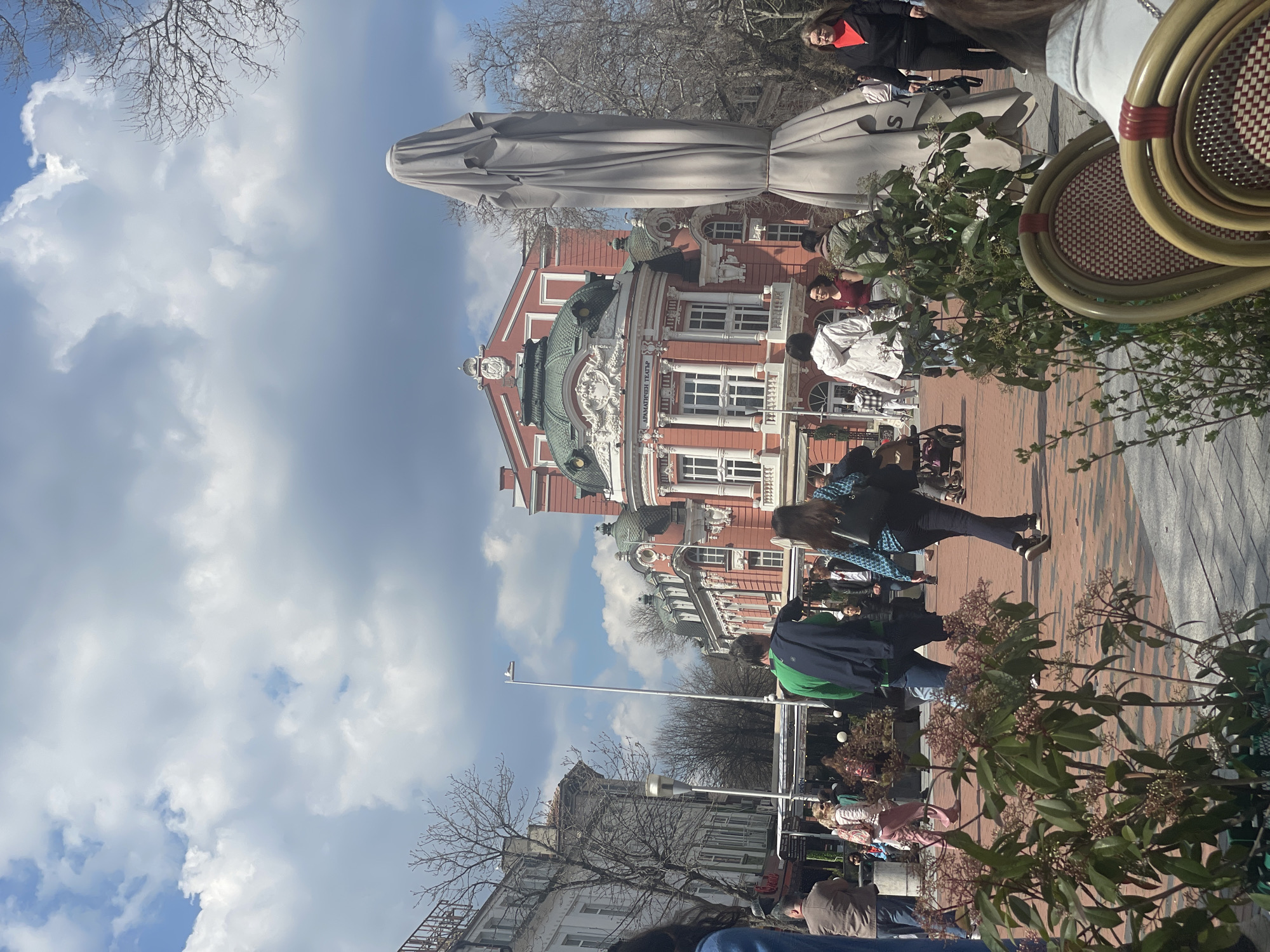 The image shows a street scene in Varna, Bulgaria, in front of the Ivan Vazov National Theatre. The theater building, with its distinctive red and white facade and green dome, is a prominent architectural feature. The sky is partly cloudy, with patches of blue visible.
In the foreground, people are walking along a brick-paved pedestrian area, some appearing to be tourists. Outdoor seating areas with tables and chairs are visible, suggesting cafes or restaurants. Foliage from potted plants or shrubs is present. A large umbrella provides shade over a table. Trees with bare branches are present, suggesting it is not summertime.