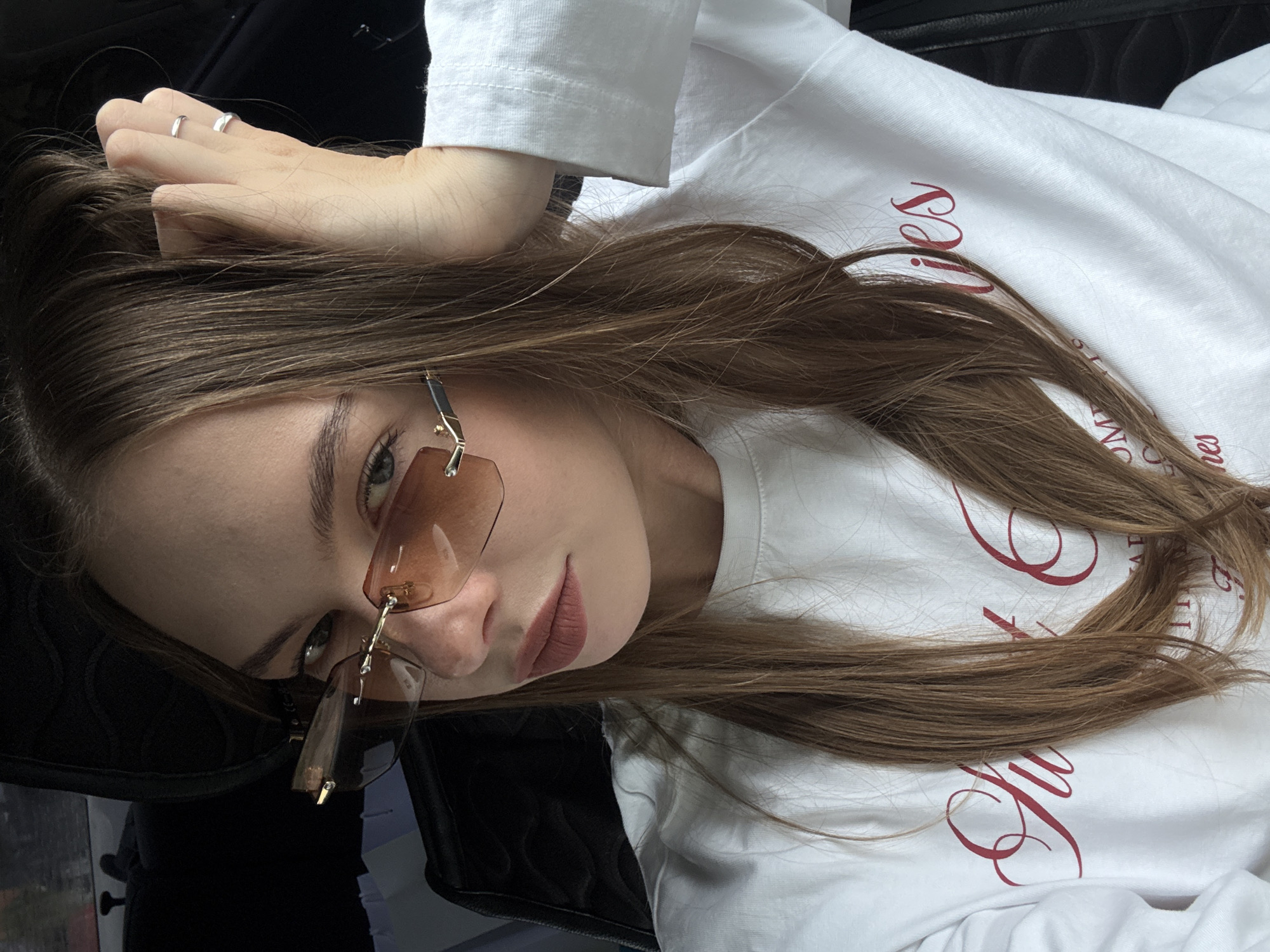 A young woman with long, brown hair is sitting in the driver's seat of a car. She is wearing a white long-sleeved shirt with red text that reads "Sweet Lies." She has on rimless, brown-tinted sunglasses and a silver ring on her left index finger. Her right hand is resting on the back of her head, pulling some of her hair. The interior of the car is dark, with the headrest visible behind her. The lighting suggests it is daytime, possibly overcast, as there is no direct sunlight. The image is a close-up, focusing on the woman's face and upper body. This scene is in the city of Perm, Russia.