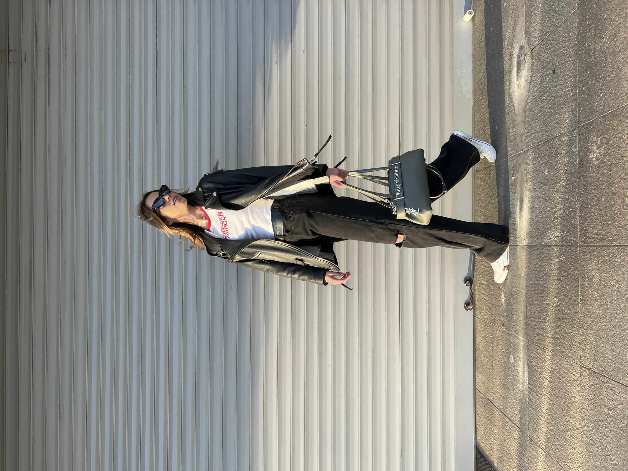 A young woman stands on a grey sidewalk next to a grey roll-up door in an outdoor setting, likely in Bar, Montenegro, based on the request. She wears black denim jeans with a rip at the knee, a white t-shirt with red lettering, a black leather jacket, and white sneakers with red detailing. She carries a grey "Juicy Couture" handbag. The background consists of the grey roll-up door on one side and the grey sidewalk and some architectural element on the other. The lighting suggests late afternoon or early morning sunlight.