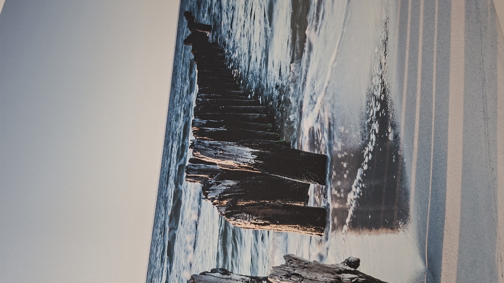 The image depicts a weathered wooden breakwater extending into the ocean under a clear sky. The breakwater is composed of numerous vertical wooden posts, some of which are partially submerged in the water, and show signs of age and erosion. Waves gently lap at the shore, creating a foamy white line where the water meets the sand. The sand in the foreground is wet and reflective, with faint, parallel shadows cast across it. The overall scene is serene and natural, capturing the raw beauty of a coastal landscape. There are no people or man-made structures visible beyond the breakwater itself. The image does not depict any location in the city of Podgorica, Montenegro, as Podgorica is an inland city and not situated on the coast.