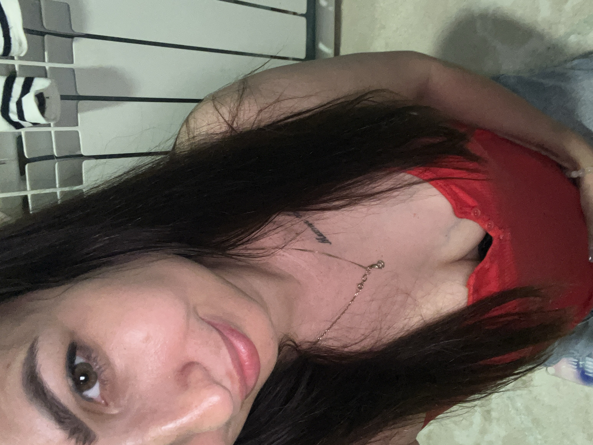 The media file is a close-up, rotated portrait of a woman. She has long, dark hair that falls across her face and chest. Her skin appears fair, and she is wearing a red top. A delicate necklace with a small pendant is visible on her collarbone, and there's a tattoo on her neck. Her eye, with a brown iris, is visible, as is her smiling mouth. The background appears to be a tiled surface, possibly a bathroom wall, with a rack holding items with black and white stripes. The lighting suggests an indoor setting, possibly during the day due to the natural light illuminating her face. The overall mood is relaxed and casual.