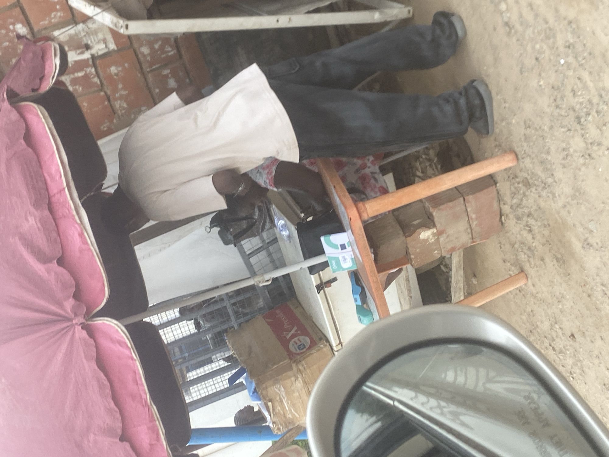 The image depicts a street scene in Iddo Tudunwada, Nigeria, under a large, pink umbrella. A person, seen from behind, is wearing a light-colored t-shirt and dark pants. They are standing near a small orange table, which is set up with various items, including what appear to be packaged goods, a blue item that might be a stapler, and a small black bag. Behind the table, a cardboard box with "X-press" branding is visible. To the left, a car's side mirror is partially visible, reflecting some of the scene. The background shows a brick wall and some metal fencing. The ground is dusty earth.