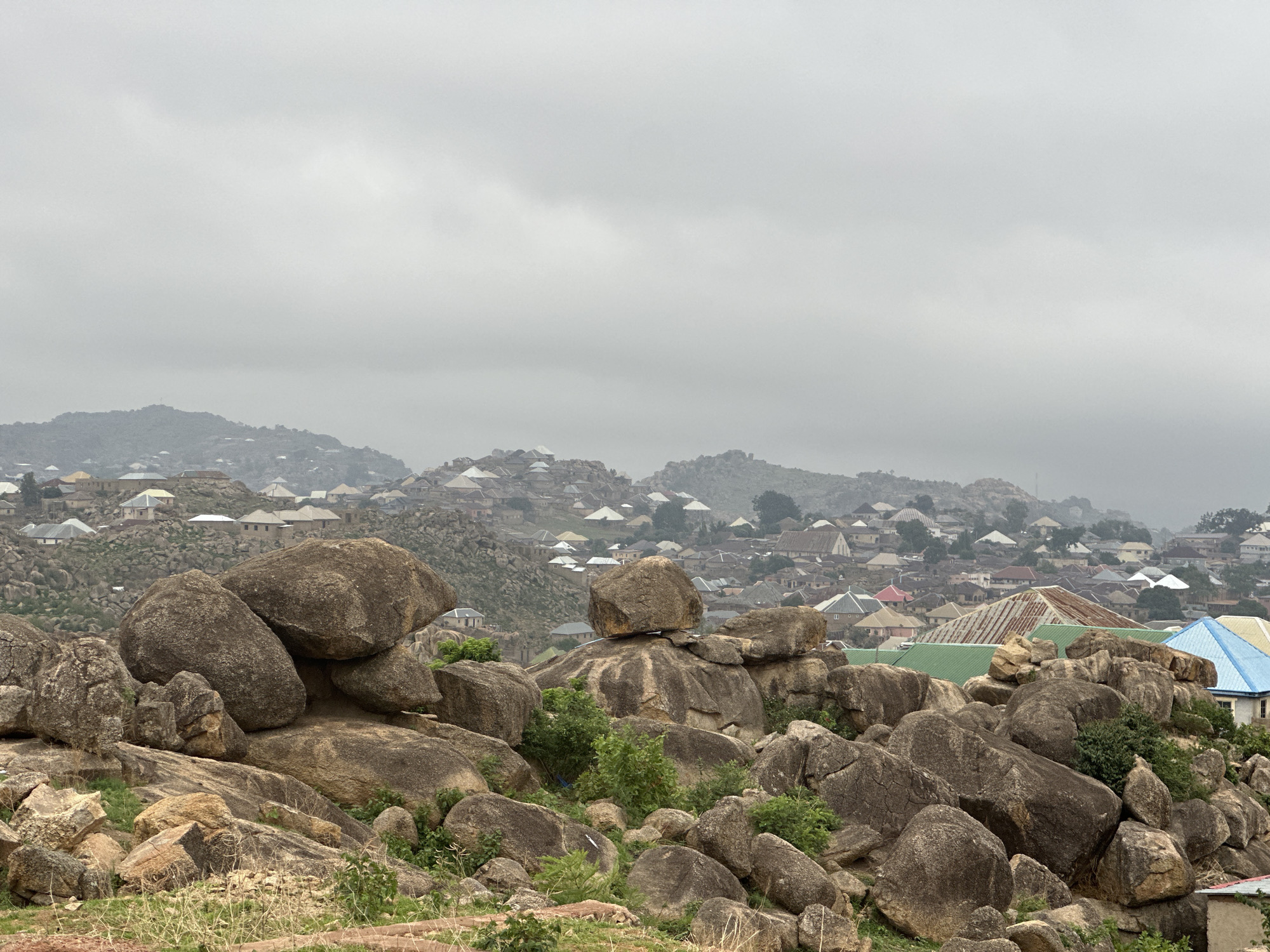 The image shows a landscape near Jos, Nigeria, characterized by large, dark gray and brown rocks in the foreground. These rocks are irregularly shaped and piled atop each other. Between the rocks, there is some green vegetation and low-lying grass. The middle ground reveals a dense urban area with closely packed buildings, many of which have white walls and brown or tan roofs. The buildings are situated on an elevated and undulating terrain, possibly a hillside. The background is hazy due to the overcast sky, and further elevated areas are partially obscured by mist or haze. The weather appears to be cloudy and the lighting is dim. There are no visible people or animals.