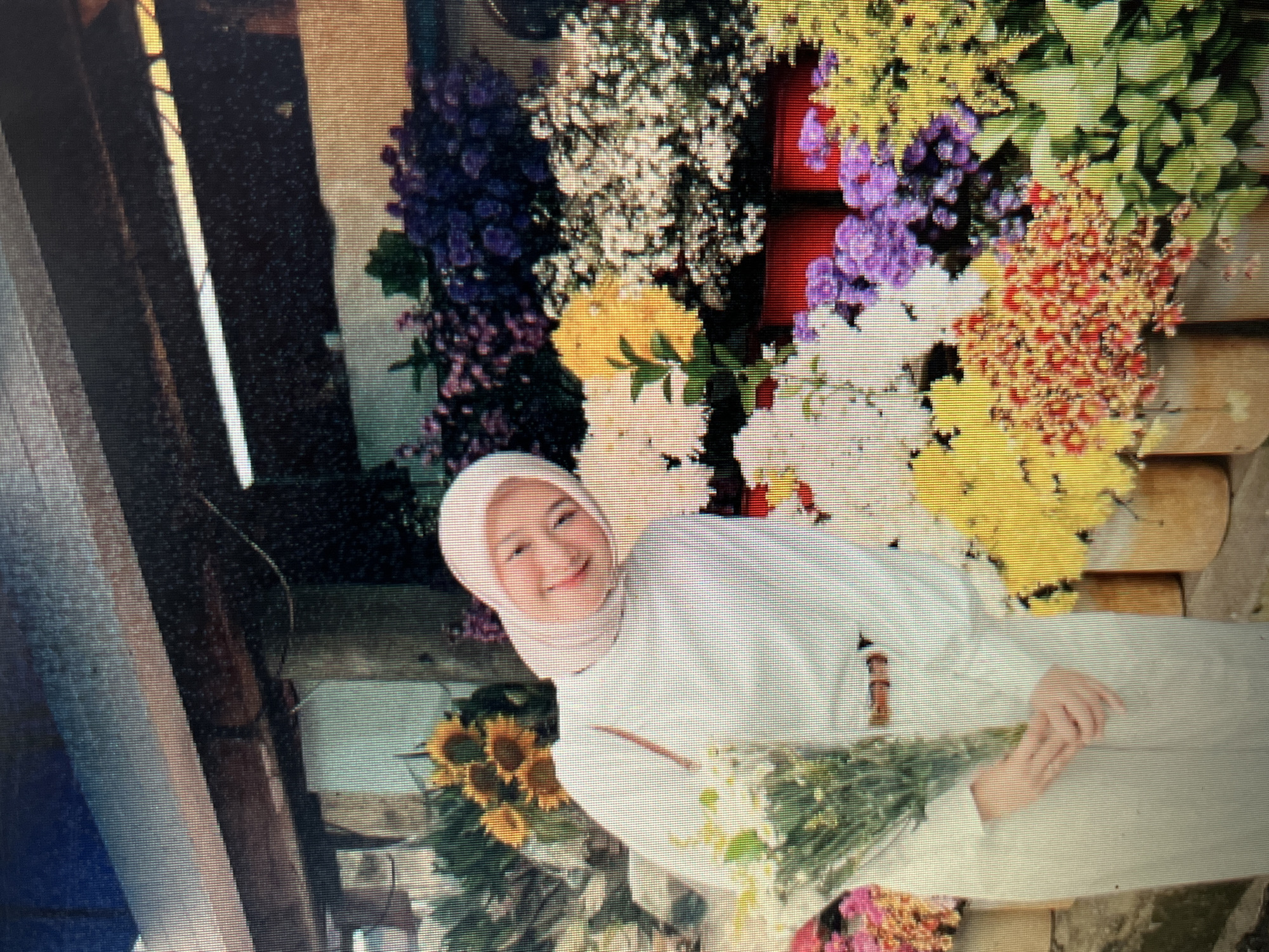 A young Indonesian woman wearing a light pink hijab and a matching white outfit stands in front of a flower stall. She is holding a bouquet of white flowers in her left hand and is smiling at the camera. The stall is filled with a variety of colorful flowers, including sunflowers, purple asters, yellow daisies, and red blossoms. The background shows a rustic wooden structure, possibly part of a market stall or an outdoor shop. The overall scene suggests a vibrant and lively atmosphere, likely in a marketplace within the city of Sejahtera, Indonesia.