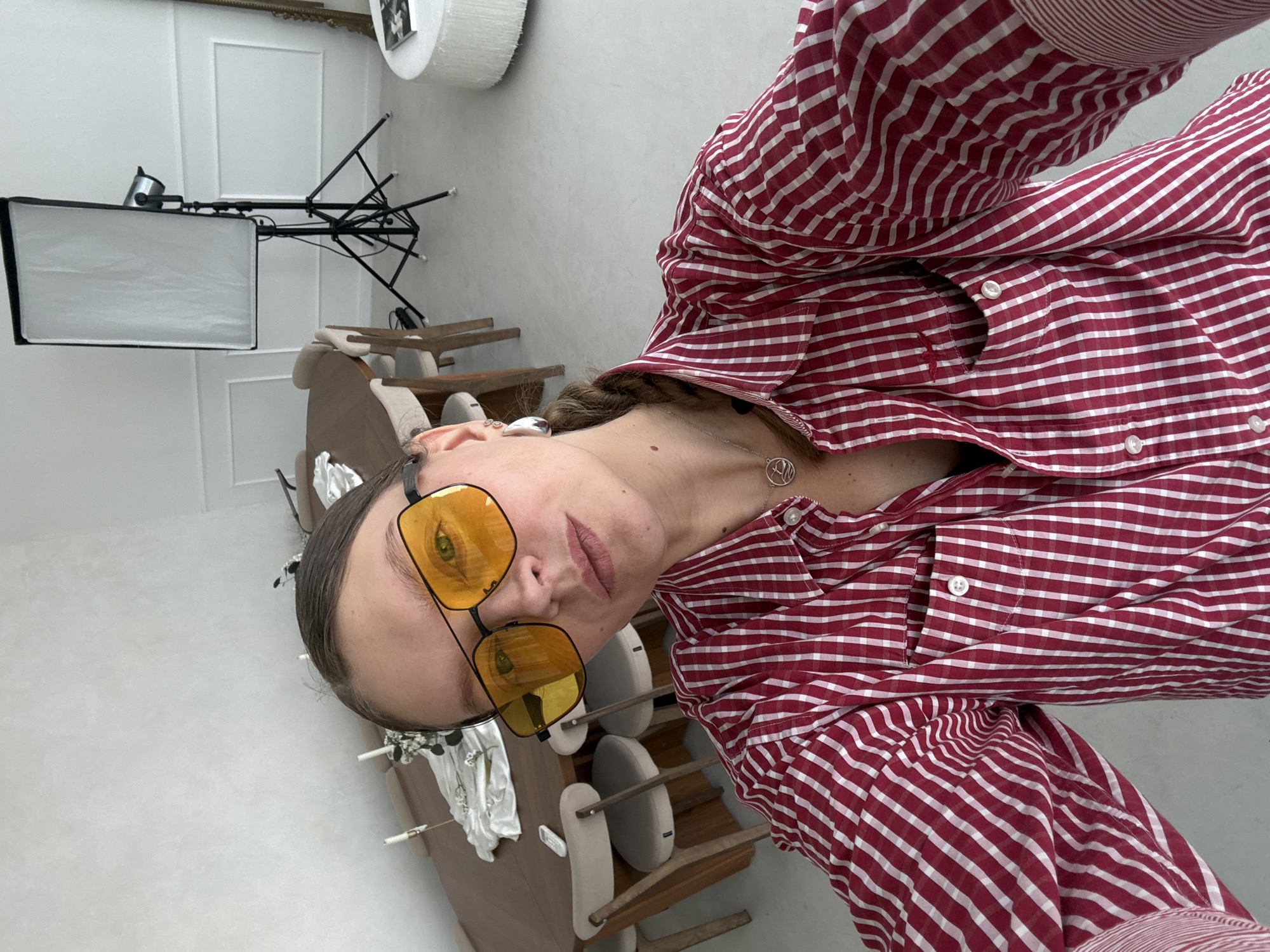 A woman with braided hair and yellow sunglasses is taking a selfie in a bright, modern room. She is wearing a red and white striped button-down shirt. The room appears to be a living or dining area with a large oval table and chairs in the background. A photography light stand is visible on the right side of the image. The floor is a light, neutral color. There is no visible indication of the city of Izhevsk, Russia in the image.