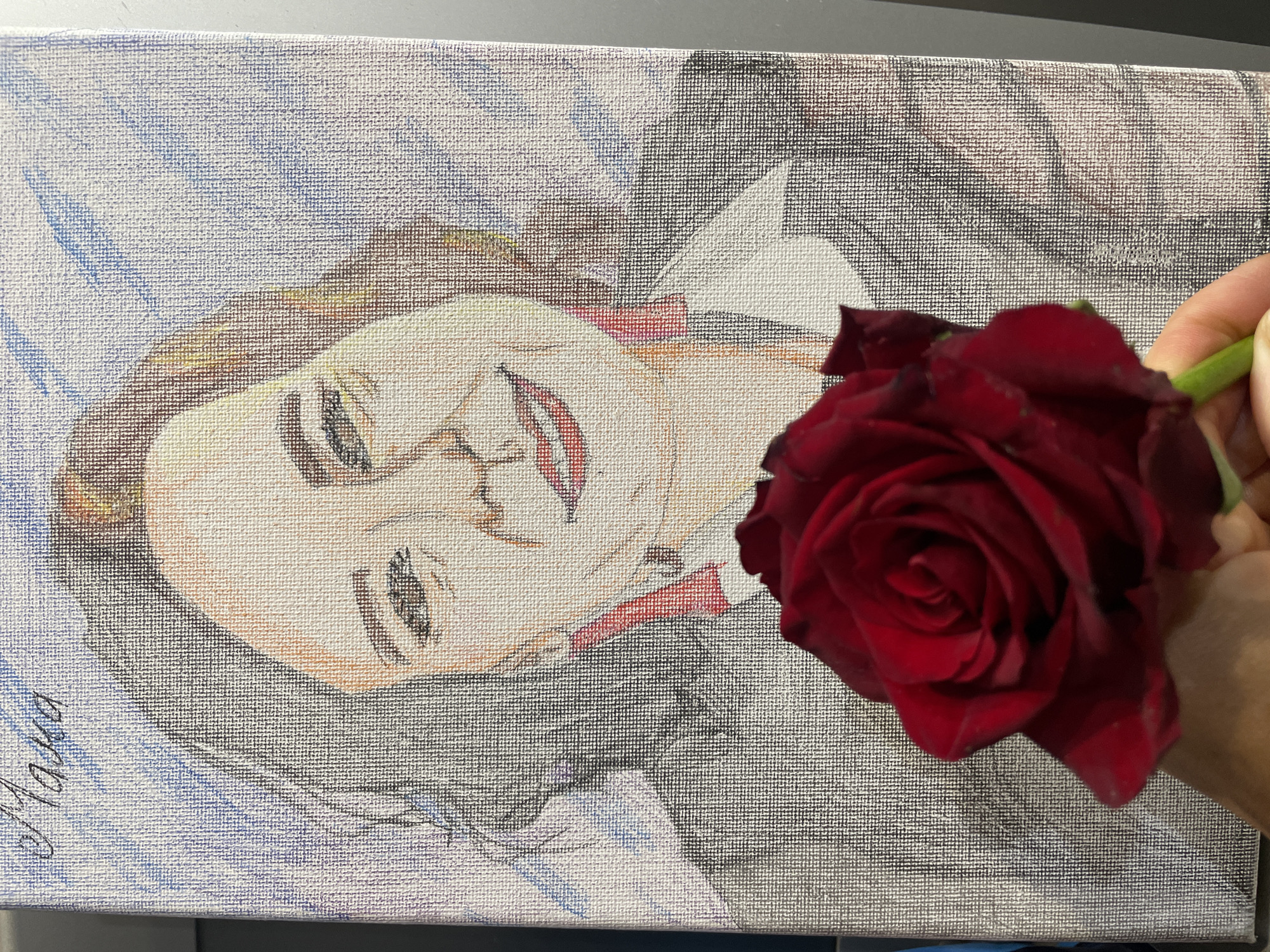 A canvas painting of a person is held in the lower left corner. A deep red rose is held in a person's right hand, partly obscuring the painting. The person in the painting is lying down with their face up and smiling. The background of the painting is a mix of blue, white, and grey tones. The canvas appears to be of textured material. The location of Sochi, Russia, is not evident.