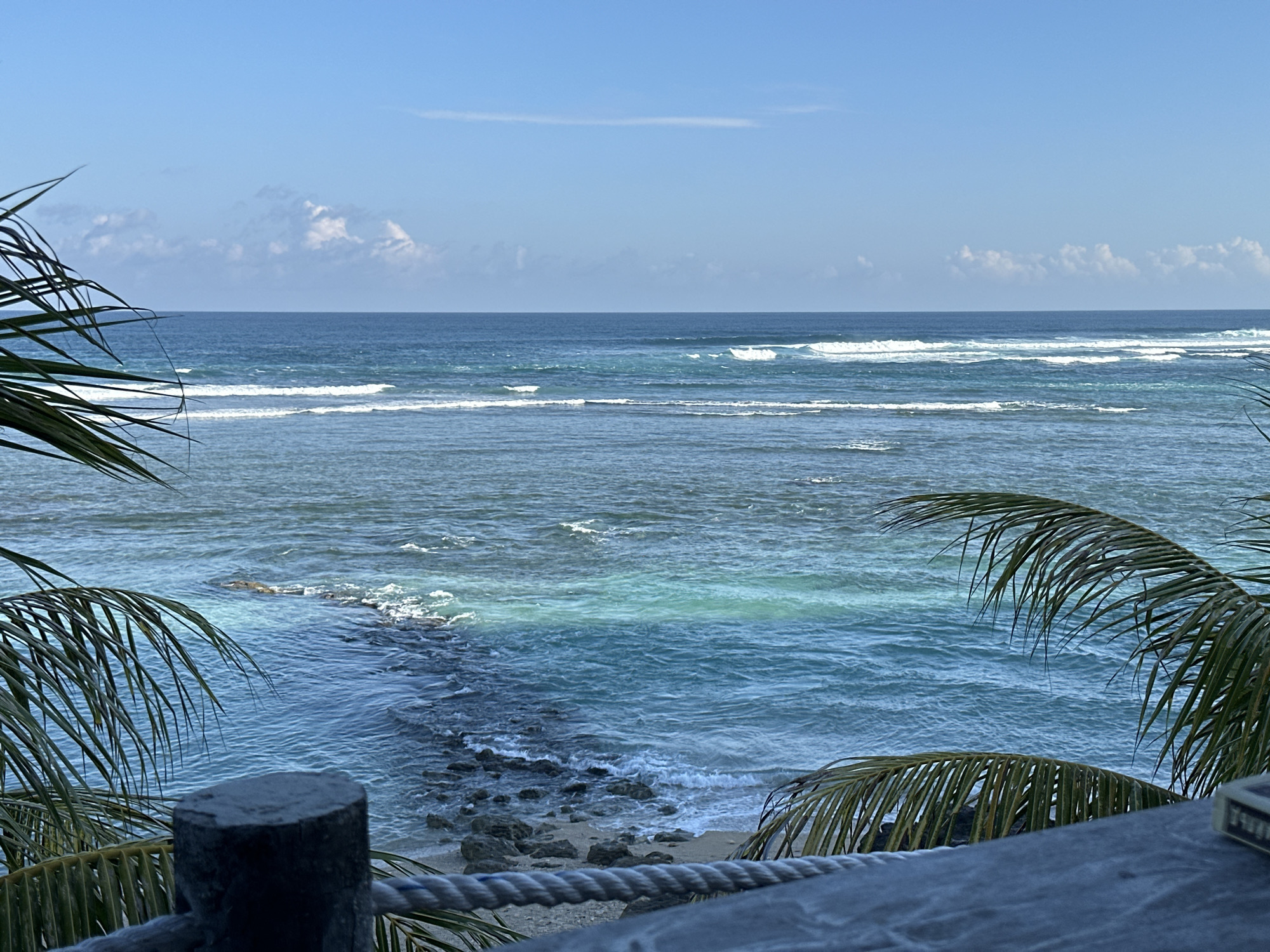 This image depicts a serene ocean view in Kuta Selatan, Indonesia. The scene is dominated by the vast blue ocean, with gentle waves rolling towards a rocky shore. The water exhibits a gradient of blues, from a deep sapphire near the horizon to a lighter, translucent turquoise closer to the shore, revealing the seabed. A prominent rocky formation juts out into the water, breaking the waves into white foam. Palm fronds frame the foreground on both the left and right sides, their fronds casting shadows and adding a tropical feel. In the lower foreground, a weathered wooden post and a thick rope barrier are visible, suggesting a viewing platform or railing. The sky above is a clear, bright blue, with a few wispy white clouds scattered across it. The overall impression is one of tranquility and natural beauty, typical of coastal areas in Bali.
