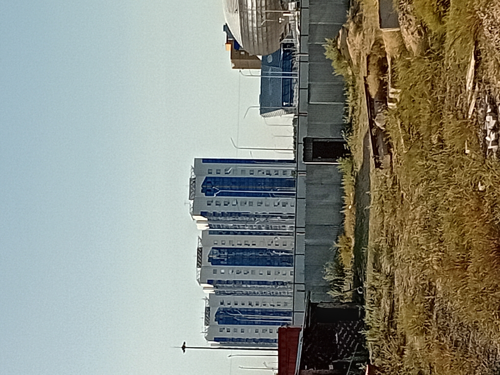 The image captures a view of modern residential buildings in Semey, Kazakhstan. In the foreground, a chain-link fence runs horizontally across the frame, with a small, dark doorway visible within it. Beyond the fence, dry, overgrown grass and a few scattered construction materials, including wooden planks and concrete blocks, are present. The main focus of the image is a row of three identical, tall, multi-story apartment buildings with a distinctive architectural design featuring white and blue facades and numerous windows. In the background, other structures are visible, including a large, domed building with a metallic finish and a smaller, angular, dark-colored building. The sky is clear and light blue, indicating daylight. The overall scene suggests an area undergoing development or situated on the outskirts of the city, with a mix of ongoing construction and completed modern architecture.