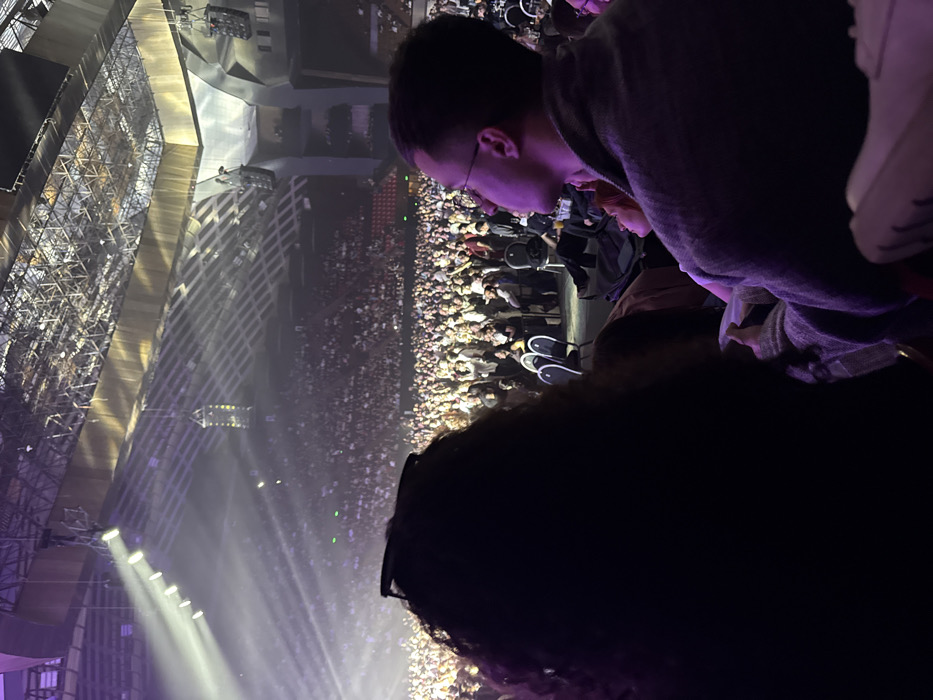 The image captures an indoor concert or event within the Karen Demirchyan Complex (Hamalir) in Yerevan, Armenia. The perspective is from within the audience, looking towards the stage area and a large crowd.
In the foreground, the back of a person's head with dark, curly hair is visible on the left, with sunglasses resting on top. To the right, a male in glasses and a dark grey jacket is seen in profile, looking slightly upwards and to the right. A portion of a female's face with reddish hair, smiling upwards, is visible between these two individuals, slightly below the male's chin. Indistinct dark seating and a white shoe are partially visible at the bottom of the frame.
The midground is dominated by a dense crowd of people, occupying the floor area and the lower tiers of stadium-style seating. Many individuals are standing, some with their arms raised, indicating active engagement with the event. Empty black chairs are interspersed in some audience areas.
The background reveals the expansive stage on the far left, intensely illuminated by multiple bright white spotlights projecting beams of light across the arena. The stage itself and any performers are largely obscured by light and distance. Above the crowd, the distinctive, intricate metal truss ceiling structure of the Karen Demirchyan Complex is visible, along with suspended speaker arrays or lighting rigs. The arena walls feature angular, geometric paneling. The overall environment is dimly lit by ambient purple-tinged lighting, contrasting sharply with the bright stage lights.
