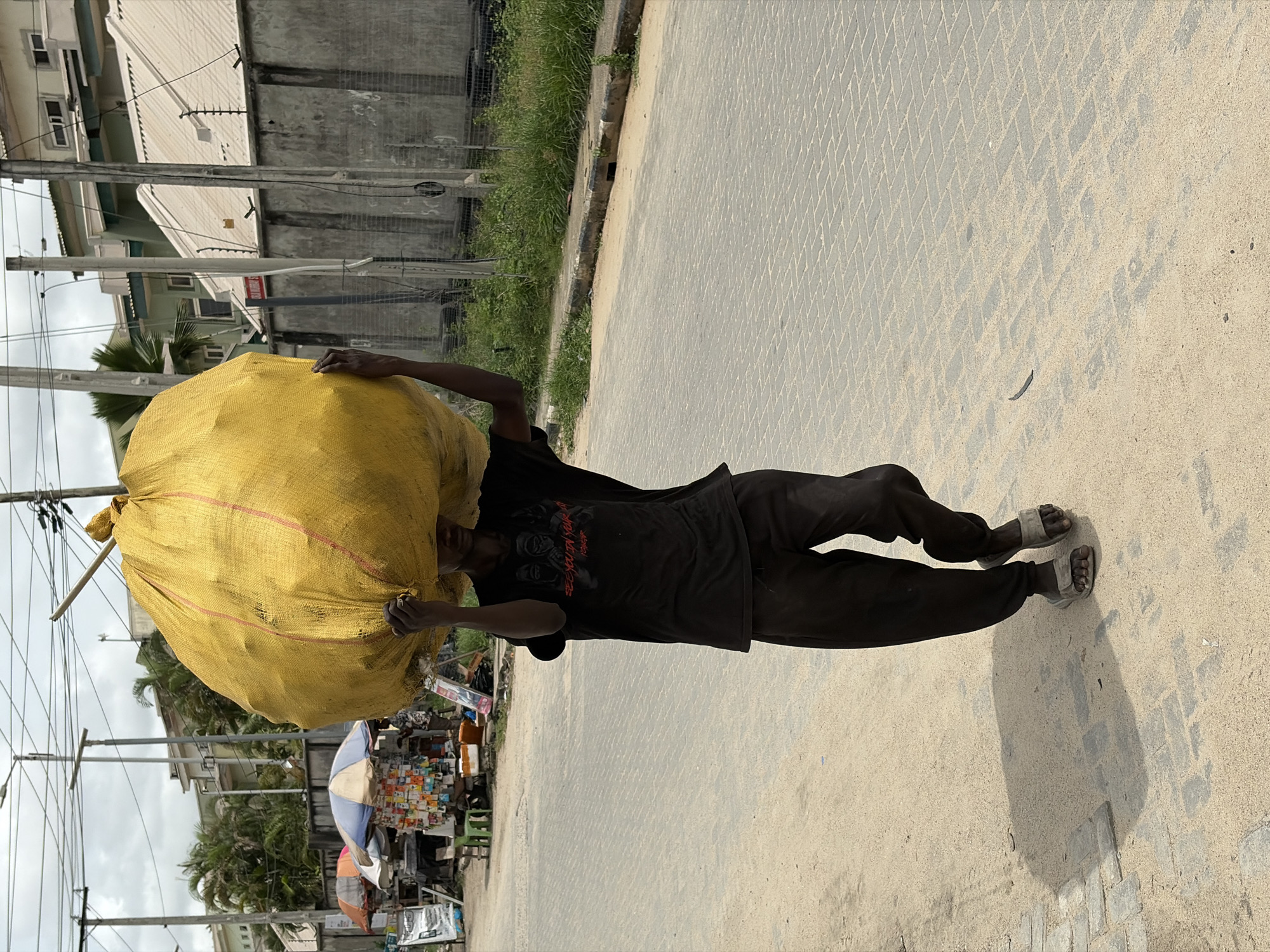 An outdoor, brightly lit image captures a person, likely a man, walking while carrying a substantial, bulging yellow sack on their head. The person is dressed in a black t-shirt, black trousers, and light-colored sandals, using their hands to stabilize the large load. Red text is visible on the t-shirt, reading "WAY YOU DO" above what appears to be "FOREVER."
The individual is on a paved surface featuring a distinct diamond-shaped pattern. To their right, this paved area transitions into an unpaved strip with green grass, behind which utility poles with numerous electrical wires stand. In the background, several buildings of varying sizes, possibly residential or commercial, are visible. Further along the street, a small market stall is set up, complete with an umbrella and displaying a diverse array of packaged goods.
The strong sunlight and clear, well-defined shadows cast by the person and the utility poles suggest it is midday or early afternoon under a clear sky. The overall scene, including the architecture, street vending, and method of transport, aligns with an urban or semi-urban setting in Igboefon, Nigeria.