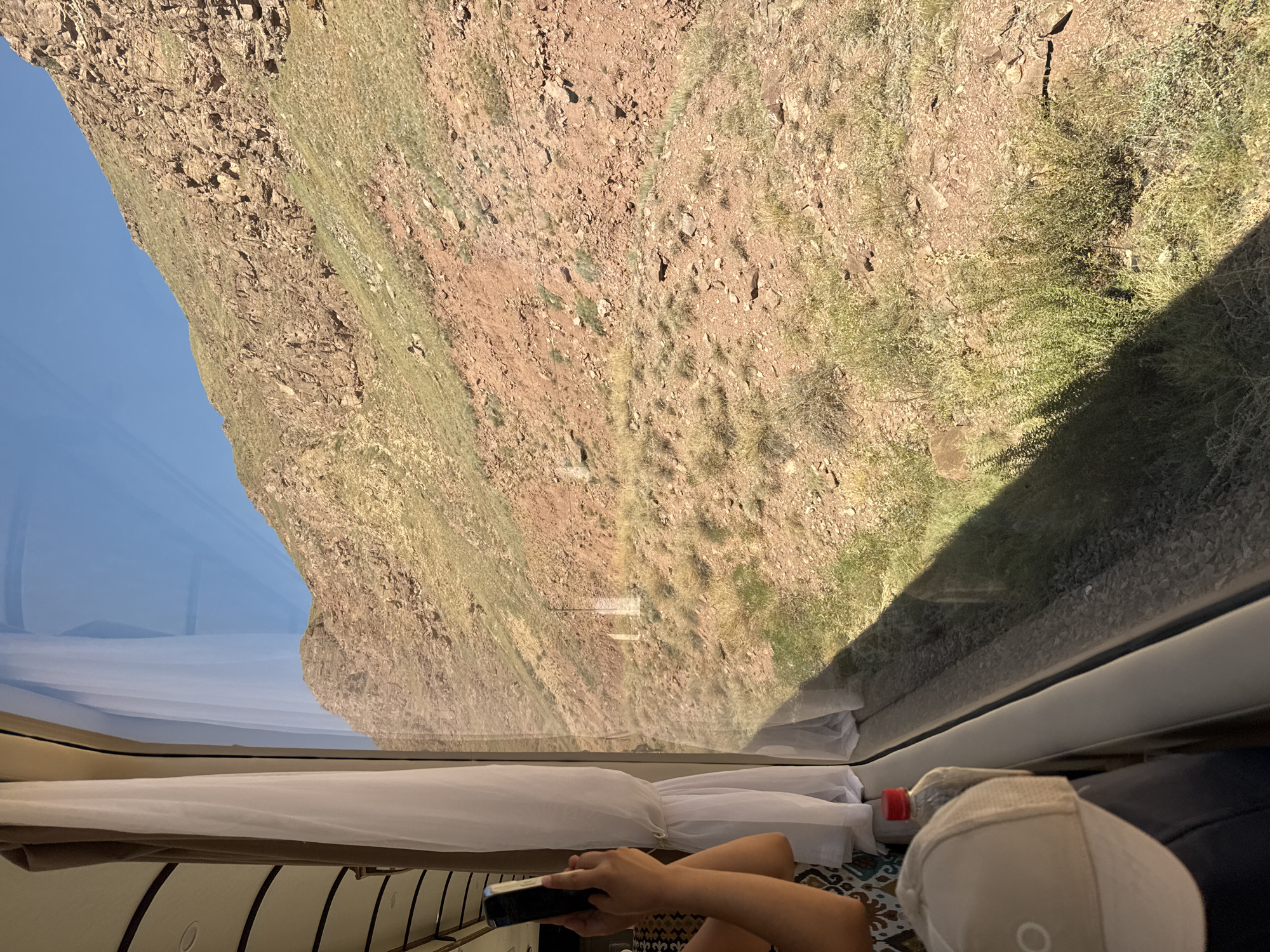 This image shows a view from inside a train, looking out at a mountainous landscape. A person's hands are visible on the left, holding a smartphone. A white curtain is drawn back to reveal the window. The exterior scene features a steep, rocky hillside with sparse vegetation under a clear blue sky. The location is described as Krasnyy Most, Kyrgyzstan, which is a mountainous region. The sun is casting shadows on the ground outside the train.