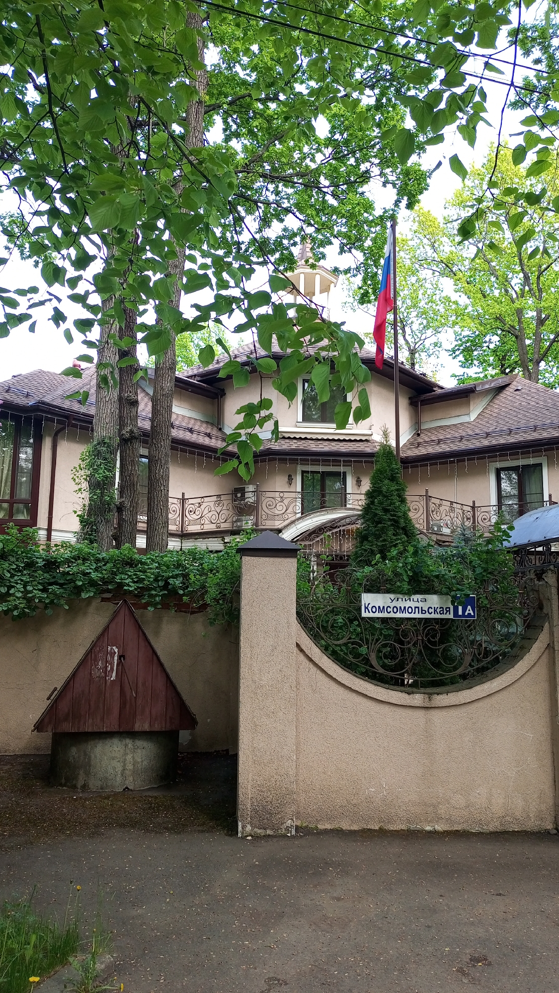 A street-level, daytime shot shows a multi-story building behind a tan textured fence with a section of decorative ironwork. The Russian flag is flying above the building. The building has a light tan exterior, a brown roof, and a small observation tower on top. A sign on the fence reads "улица Комсомольская 1А," indicating the location is on Komsomolskaya Street, building 1A. To the left of the fence is a well with a wooden A-frame roof. Trees and foliage surround the building. The image was taken in Дубки, Russia.