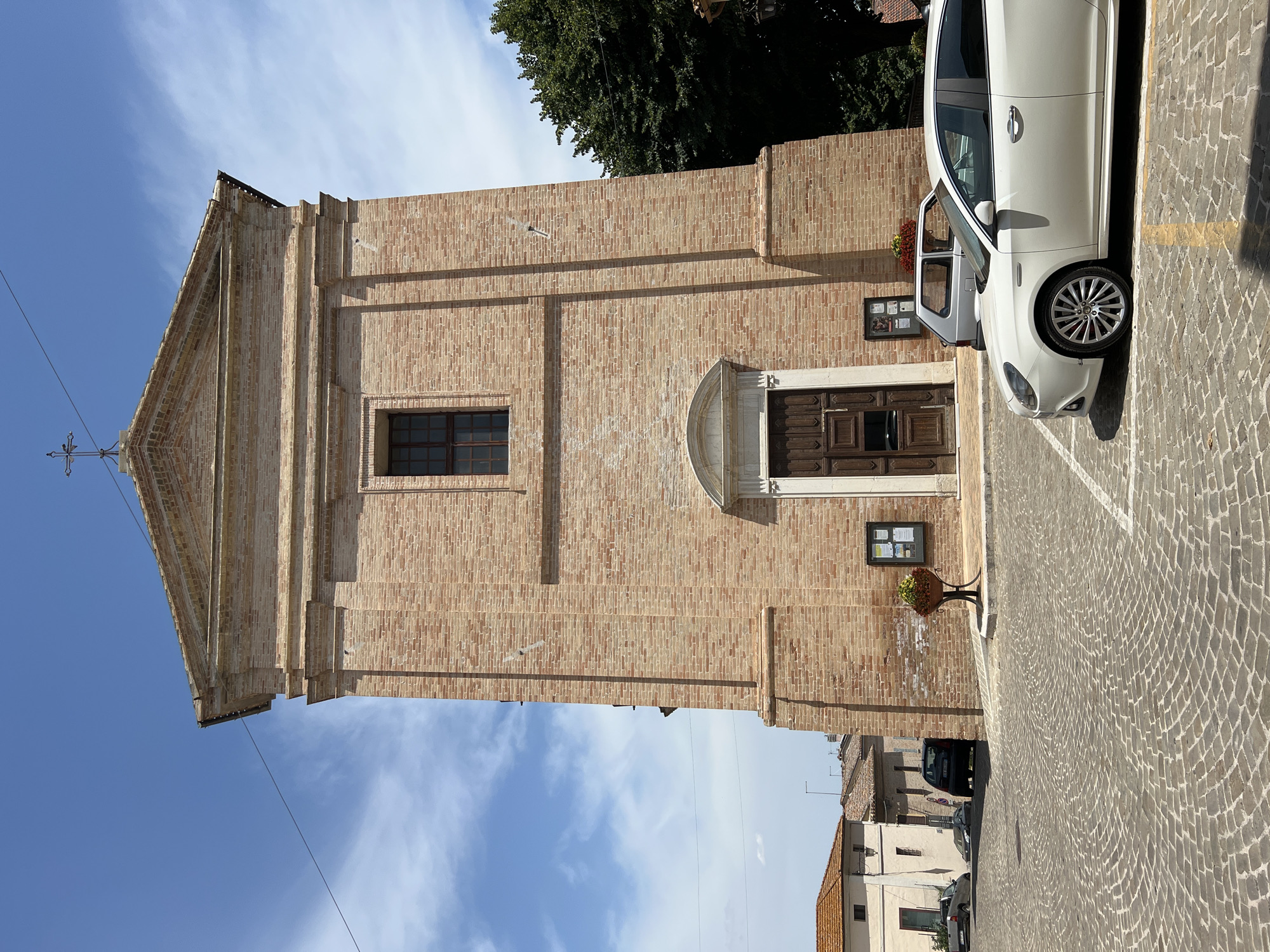 The image shows the facade of a brick church in the city of Barbara, Italy. The church is tall and narrow, with a triangular pediment at the top and a cross. The facade has a large wooden door and a single window above it. There is a potted plant with red flowers to the left of the door and a bulletin board to the right. In front of the church is a paved square with cobblestones. Several cars are parked in the square, including a white Alfa Romeo in the foreground. The sky is blue with some white clouds, and a few trees are visible to the right of the church. The overall impression is a sunny day in a quiet Italian town.