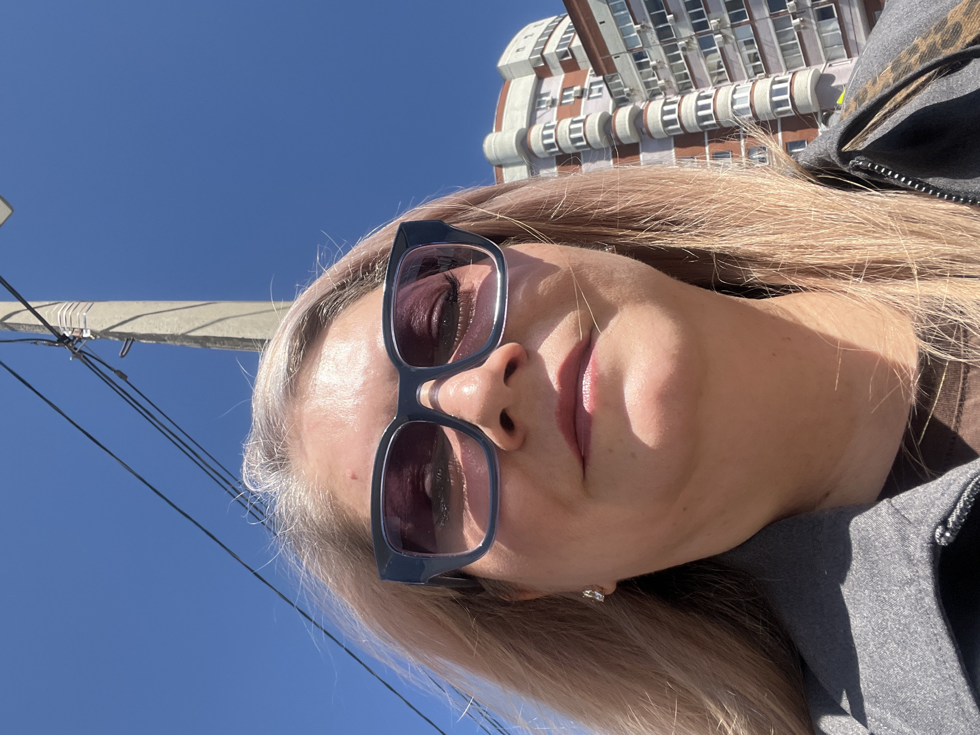 A woman with blonde hair and sunglasses is taking a selfie outdoors on a sunny day. A utility pole and power lines are visible in the background against a clear blue sky. A building with a unique architectural design is also in the background, identifying the location as Izhevsk, Russia. The woman is wearing a gray jacket. The photo is angled upwards towards the sky.