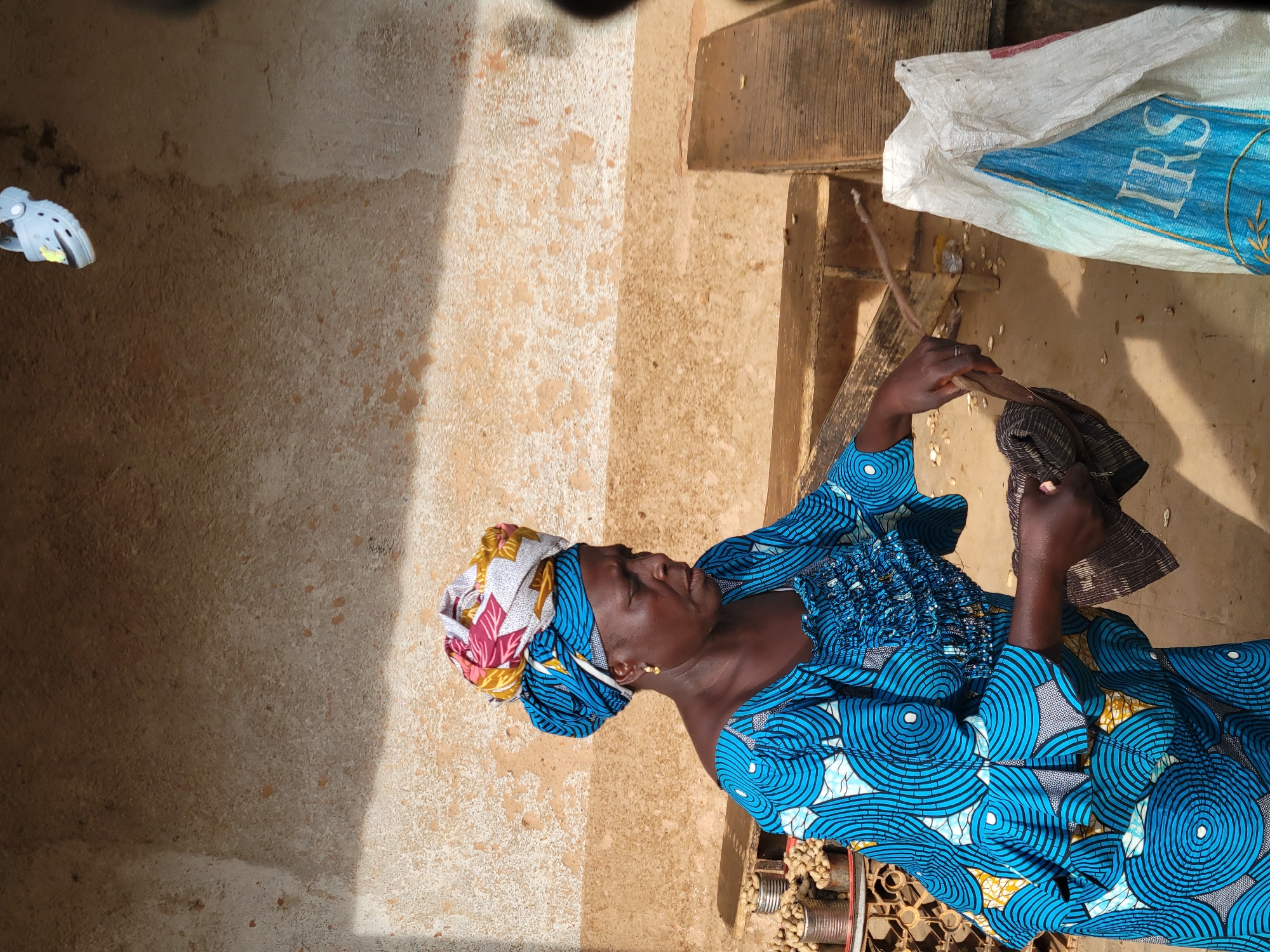 A woman wearing a blue and white patterned dress and a colorful headwrap is standing in front of a textured, light-colored wall. She is holding a dark cloth and a wooden stick, with her hands appearing to be in motion. To her left, several cans are filled with peanuts. In the foreground and to the right, a white sack with blue lettering, including the letters "IRS," is visible. The scene is outdoors, with shadows cast on the wall, suggesting daylight. The overall environment appears rustic. The image does not provide specific details that would place the location in the city of Jos, Nigeria, beyond the general visual cues of an outdoor market or street scene.