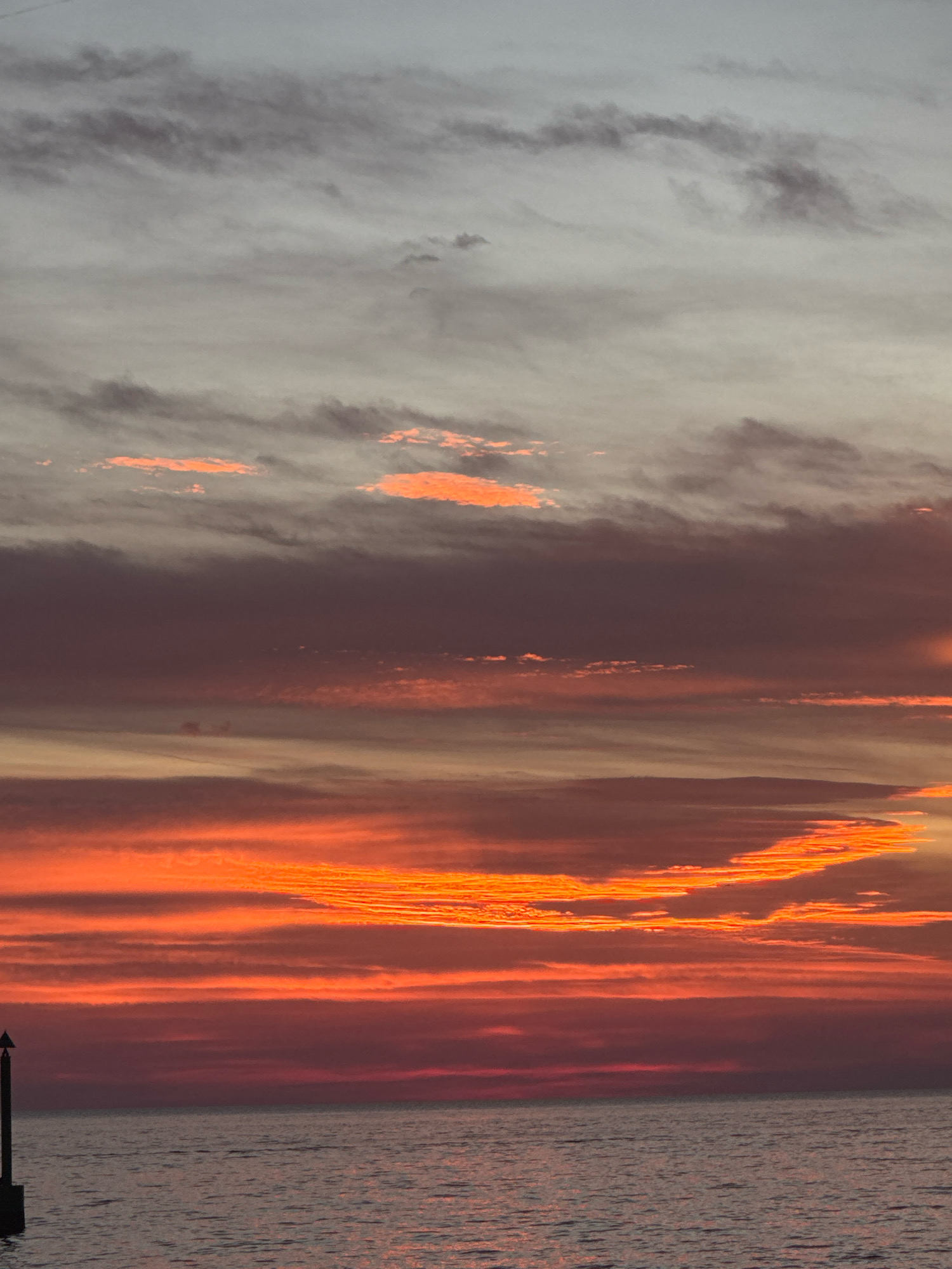 The image captures a dramatic sunset over the sea. The sky is filled with layers of clouds, transitioning from a soft gray at the top to vibrant oranges and reds closer to the horizon. The sea is a muted gray-blue, reflecting the colors of the sky. A black marker post is visible on the left side of the frame, standing in the water. The scene is devoid of any people or specific actions. The location is identified as the coast of Novigrad, Croatia.